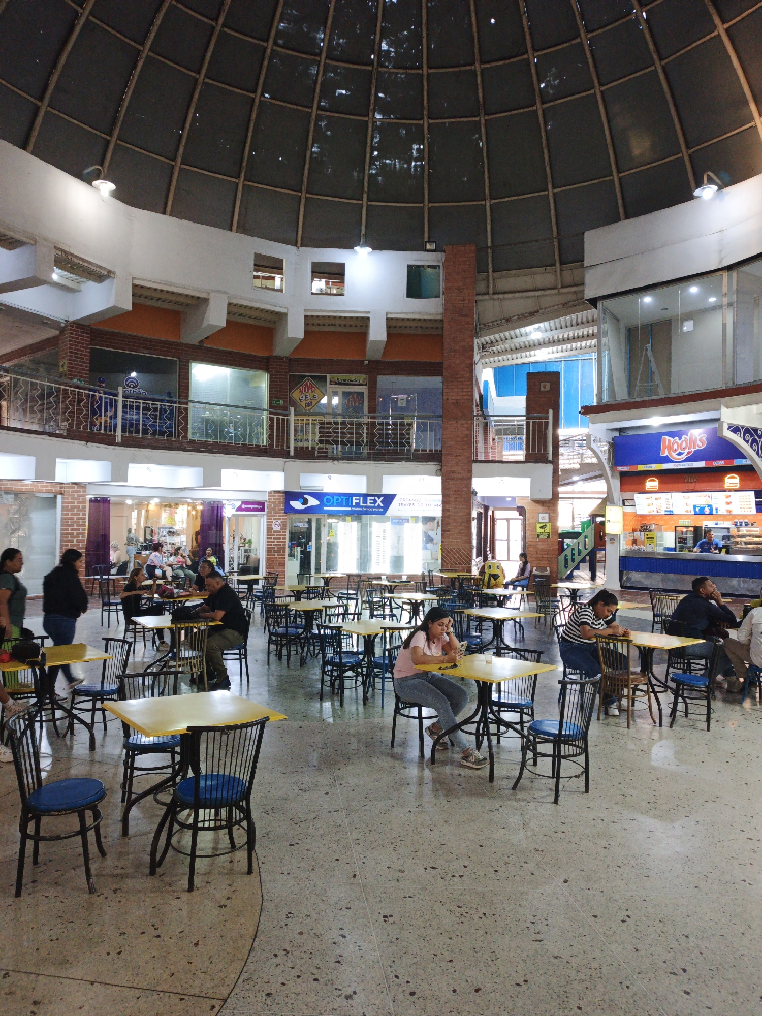 The image shows the interior of a food court in Tinaquillo, Venezuela, beneath a large domed ceiling. The space is filled with tables and chairs, occupied by people dining and using electronic devices. Food vendors are visible in the background, including one branded "Hoolis." The floor is a light, speckled terrazzo. The overall impression is of a casual, indoor gathering place.