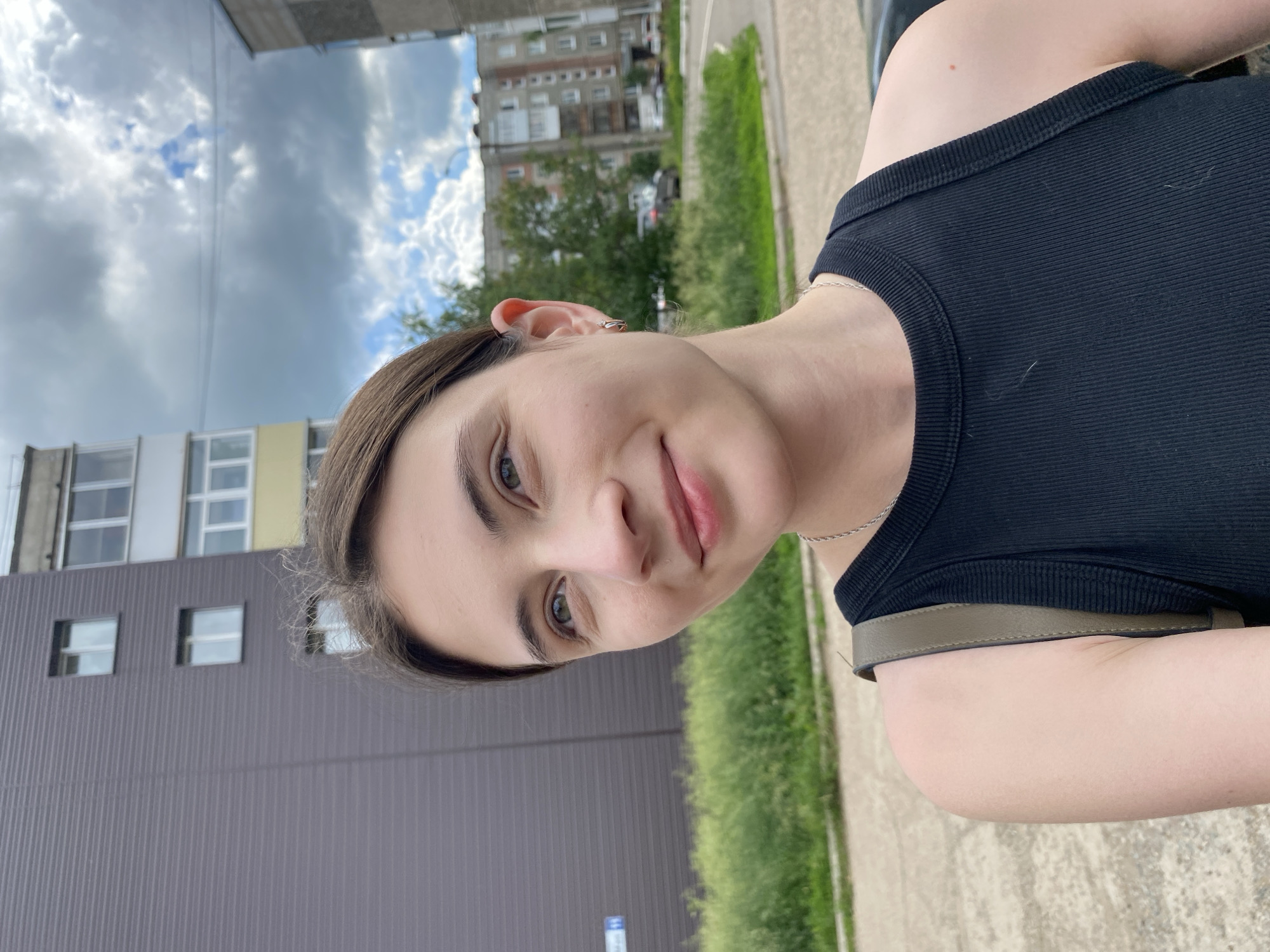 The image shows a young woman in Votkinsk, Russia. She is looking towards the camera with a slight smile. She has brown hair, brown eyes, and is wearing a black tank top. A thin silver necklace is visible around her neck, and a thin strap of a bag is visible across her shoulder. The background consists of a grey building with square windows, a green grassy area, and a cloudy sky. The time of day appears to be daytime, given the lighting. The overall scene is outdoors, in what appears to be an urban or suburban environment.