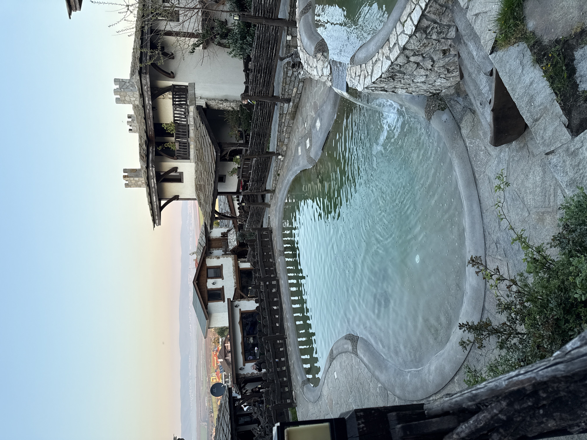 The image depicts an outdoor pool and surrounding structures in Medovo, Bulgaria. A large, irregularly shaped pool with light-colored water dominates the center. A smaller, elevated pool with a waterfall feature feeds into the larger pool. The pools are surrounded by stone and concrete.
Adjacent to the pool are buildings constructed in a rustic style, featuring white walls, dark wood accents, and stone roofing. A wooden fence borders the pool area. In the background, a valley landscape stretches towards a horizon with a gradient sky. The image is shot from an elevated perspective, possibly from a balcony or deck.