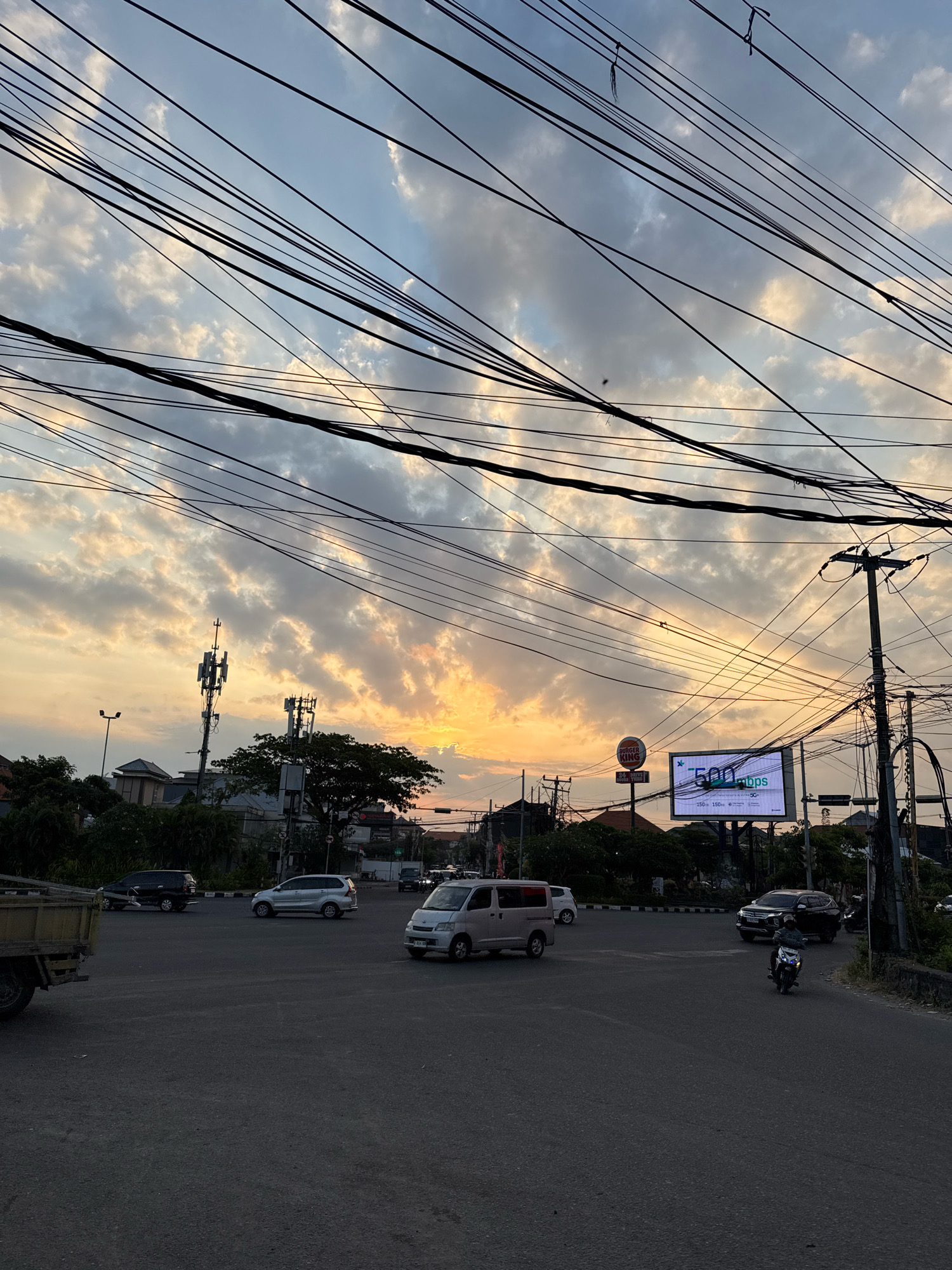 A street-level, medium shot captures a city intersection in Kuta, Indonesia, at sunset. The sky is filled with a dramatic display of clouds, illuminated by the warm hues of the setting sun. A tangled web of overhead electrical wires crisscrosses the frame, obscuring portions of the sky and the urban landscape below.
In the foreground, the asphalt road shows faint lane markings. Several vehicles are navigating the intersection: a silver SUV is turning left, a light-colored van is moving forward, and a black SUV is stopped at the intersection. A lone motorcyclist in a dark helmet and jacket is also present, approaching the intersection. To the left, the rear of a yellow flatbed truck is visible.
In the mid-ground, various buildings and infrastructure are visible. Two prominent cell phone towers stand tall, one with multiple antennas. A Burger King restaurant sign is perched atop a pole. A large digital billboard displays "500 Mbps" and promotional text related to 5G internet. Streetlights and traffic lights are also present.
The background features more buildings, trees, and the continuation of the street. The overall atmosphere suggests a busy urban environment transitioning into evening, with the natural beauty of the sunset contrasting with the man-made elements of the city.