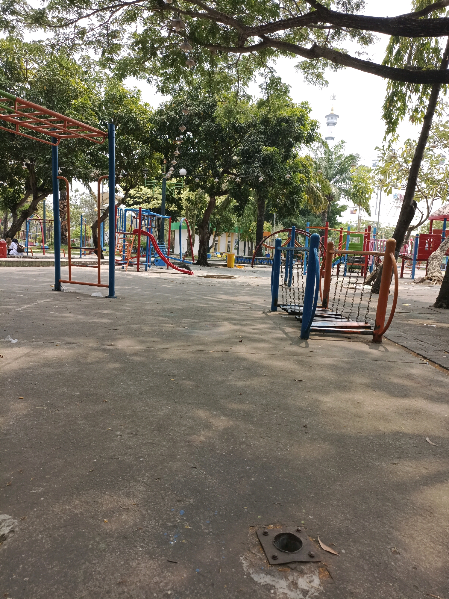 This is an outdoor photograph of a playground area, likely in Lamongan, Indonesia, as indicated by the presence of a tall tower in the background that resembles the Lamongan Tower. The playground features various colorful structures, including a red slide, monkey bars with orange and blue supports, and a chain bridge with blue and orange railings. The ground is paved with asphalt, and there are trees with lush green foliage providing shade. In the mid-ground, a few individuals are seated on a bench near the playground equipment. In the background, the Lamongan Tower stands prominently against a bright sky, and a mosque is visible near its base. The overall scene depicts a public recreational space under a bright, overcast sky.