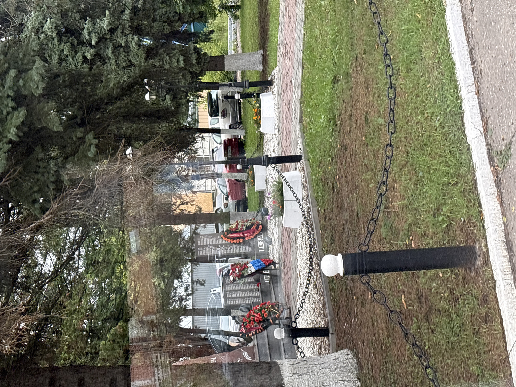 The image depicts a memorial site in Sverdlovskiy, Russia, likely commemorating fallen soldiers. A monument, possibly a wall with inscribed names or dates, is visible in the background, adorned with several wreaths of artificial flowers. Two prominent wreaths, one red and white and another featuring blue, white, and red elements (resembling the Russian flag), are placed near the monument. The area around the monument is paved with red and white bricks, and gravel pathways are visible. Two lamp posts with white spherical tops are positioned along the pathway. A chain-link fence with black posts and white caps encloses a grassy area in the foreground. Several trees, including evergreens and deciduous trees, surround the memorial. The presence of cars and what appears to be a building in the background suggests an urban or semi-urban setting. The overall scene is calm and somber, indicative of a place of remembrance.