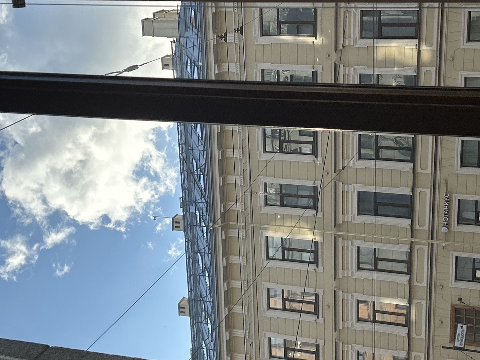 The image shows a view of an apartment building through a window with a dark frame. The building has a light yellow facade and multiple floors with numerous windows. The roof is covered with skylights and there are chimneys visible. The sky above is blue with scattered white clouds. Several wires are strung across the scene, obscuring parts of the building and the sky. A sign with Russian text, "НОТАРИУС," meaning "NOTARY," is visible on the lower part of the building. There are no people or distinct actions visible in the image. The overall scene depicts an urban street view with architectural elements of a European city.