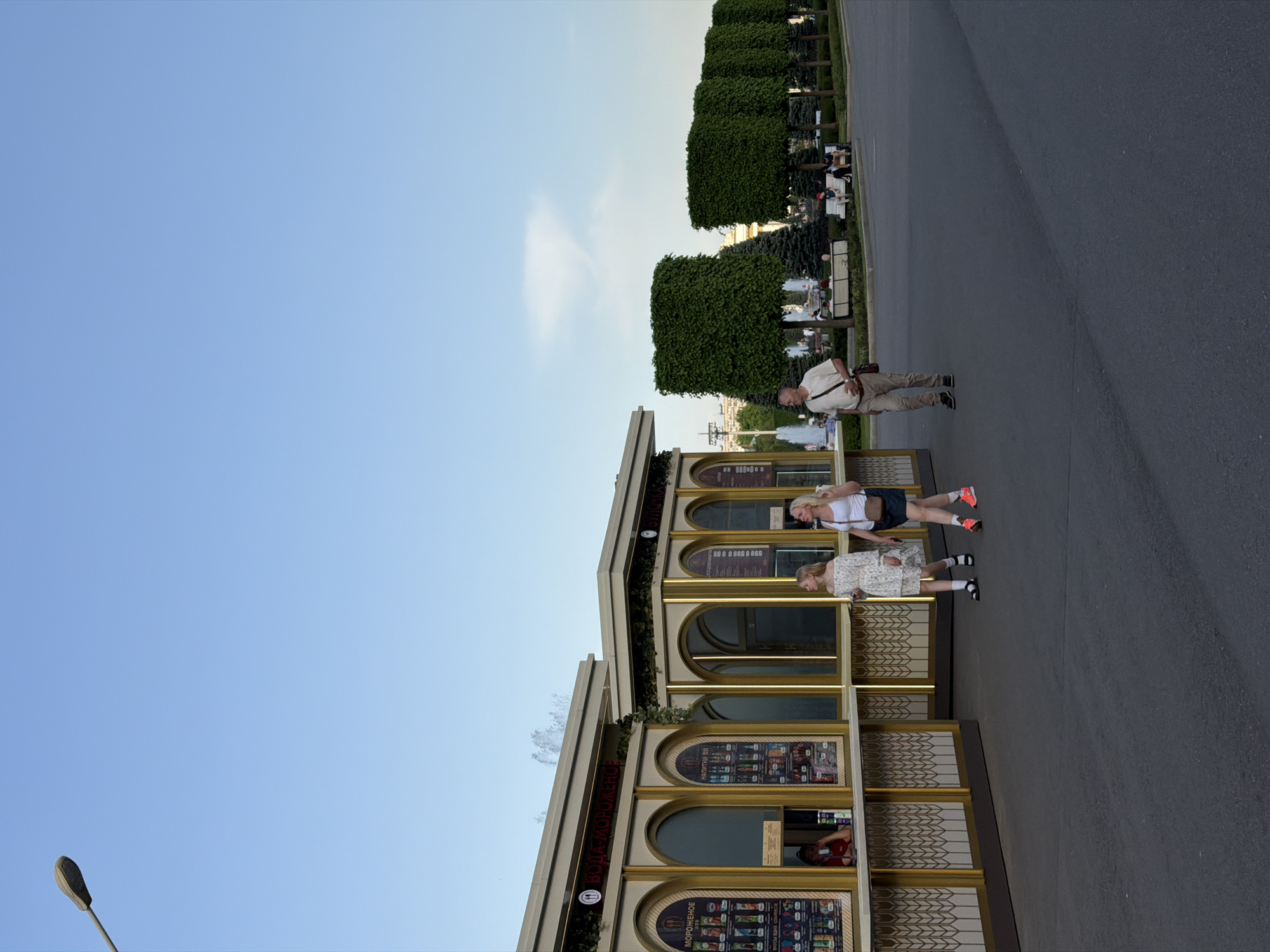 The image depicts a daytime scene in Moscow, Russia, featuring a group of people near a building with arched windows and gold detailing. Two women are standing near the building, while a man is walking on the pavement nearby. In the background, there are rows of trimmed trees and a glimpse of a fountain. The sky is blue. The building appears to be a kiosk or small shop, possibly selling refreshments. The general environment suggests an urban park or promenade.