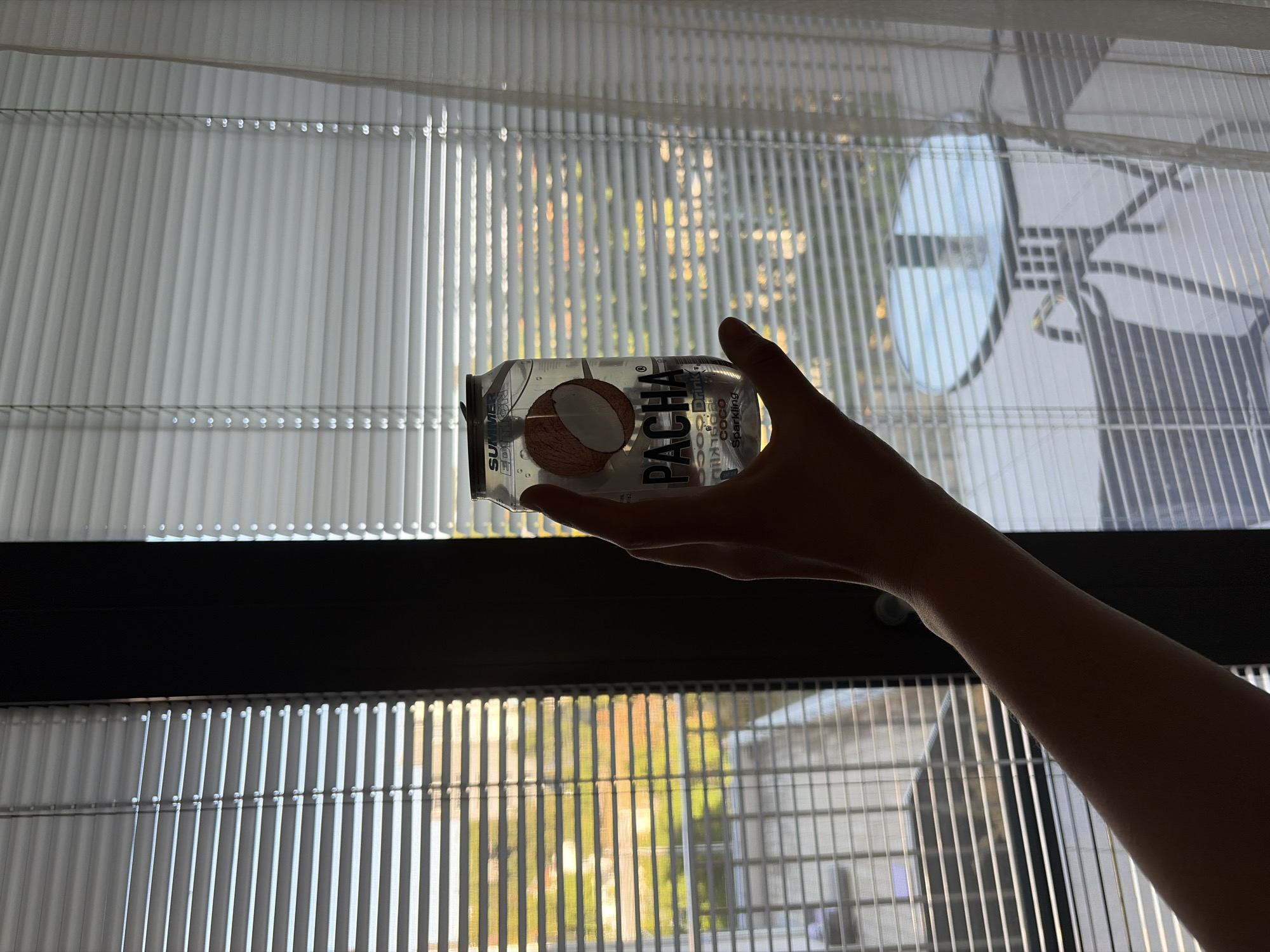 A person's hand holds a clear can of "PACHA" brand coconut sparkling drink in front of a window with horizontal blinds. The can has an image of half a coconut on it and reads "SUMMER EDITION" at the top. Outside the window, through the blinds, a blurry scene is visible showing a balcony with a table and chairs, and some greenery. The lighting suggests it is daytime, possibly late afternoon or early morning due to the soft light filtering through the blinds and the shadows. There is no information to determine the location as Nitra, Slovakia.