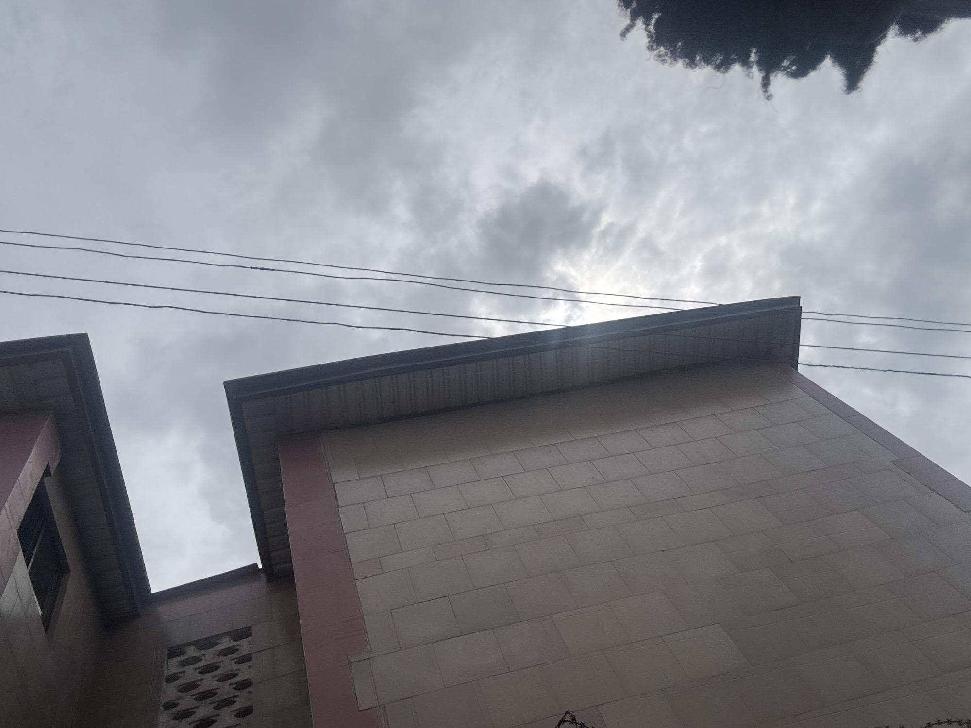 The image shows an upward angle view of a multi-story building complex in Ihiagwa, Nigeria. The buildings are constructed with a light tan brick or block and have a reddish-pink trim on the corners. The roof edges have a dark-colored overhanging fascia. The sky is heavily overcast with clouds partially obscuring the sun. Electrical wires are visible stretching across the top portion of the frame.