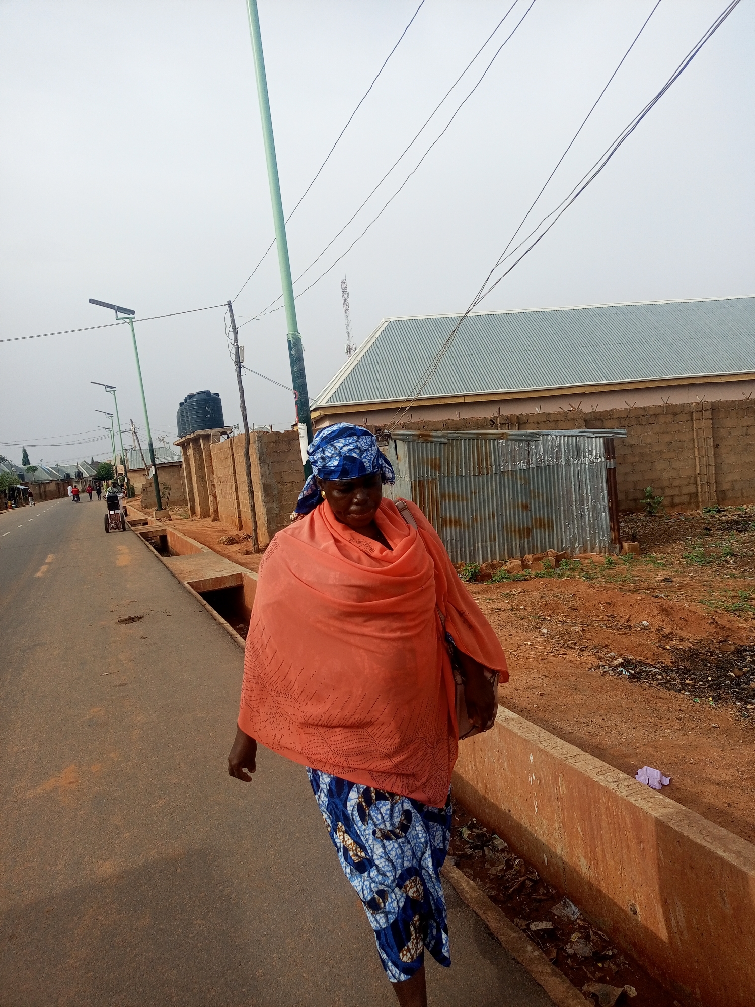 The image depicts a woman walking on a paved road in Tempure, Nigeria. She is wearing a blue patterned skirt, an orange shawl, and a blue headscarf. The road has concrete drainage ditches on the side, filled with debris. Behind the woman is a brick wall topped with barbed wire, a corrugated metal structure, and a building with a gray metal roof. In the distance, other buildings and people can be seen. The sky is overcast. A green pole stands on the roadside. Overhead wires cross the scene.