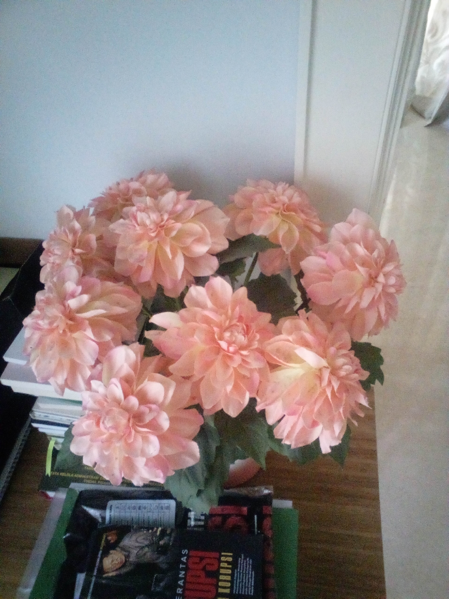 A high-angle, medium shot captures a bouquet of artificial pink dahlias in a brown pot. The flowers are arranged on top of a stack of books, some of which have visible titles in Indonesian, including "TUTUR KELOLA ADMINISTRASI" and "KORUPSI". The bouquet is positioned on a wooden table next to a white wall and a doorway. The lighting is soft and diffused, suggesting an indoor setting. No people are present in the scene. The overall impression is one of domestic tranquility. Based on the Indonesian text on the books, this scene is likely located in South Jakarta, Indonesia.