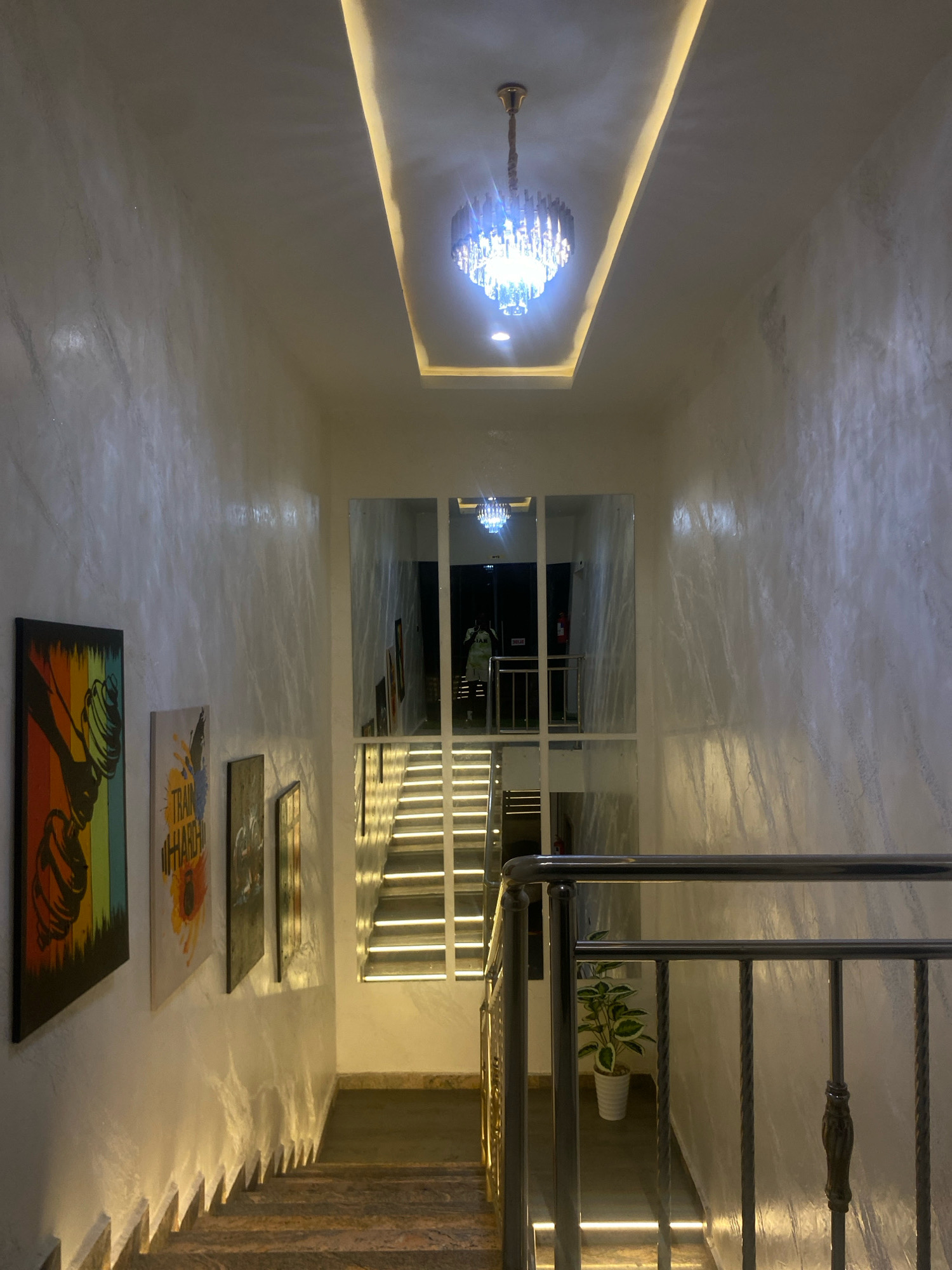 The image shows a well-lit staircase inside a building. The walls are textured with a marble-like finish and are illuminated by accent lighting along the base of the stairs. A modern chandelier hangs from the ceiling. On the left wall, several framed art pieces with motivational themes are displayed. Mirrors are strategically placed on the right wall, reflecting the staircase and the chandelier, creating an illusion of more space. A metal railing lines the stairs. A potted plant is visible on a lower level. The overall impression is one of a modern and stylish interior, possibly a hotel lobby or a commercial building. The location is stated to be Barden Yana, Nigeria. No specific people or interactions are captured in this image; it is a static scene of the interior architecture and decor.