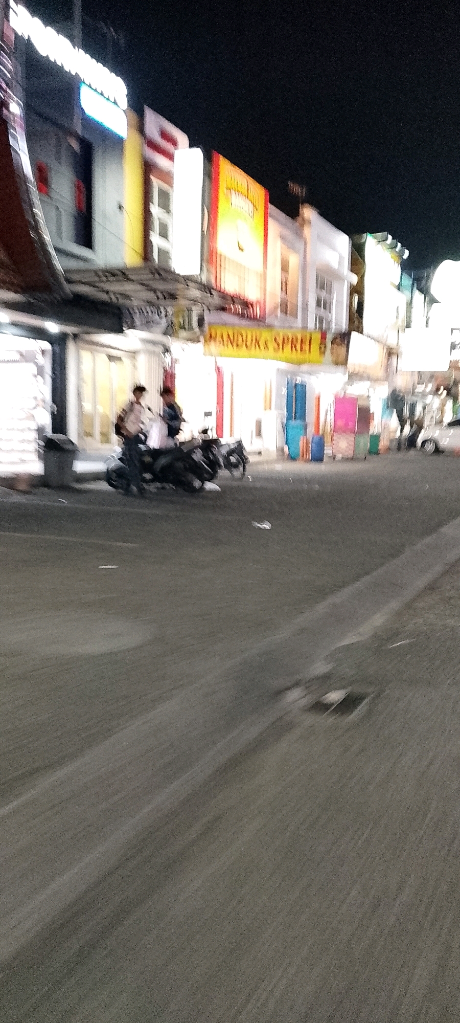 This is a slightly blurry, nighttime photograph taken from a moving vehicle on a street in Cileungsi, Indonesia. The image focuses on the storefronts lining the street, which are illuminated by neon signs and interior lighting. The signs suggest commercial activity, with one prominently displaying the text "HANDUK & SPREI" (Towels & Sheets). In the foreground, a dark-colored motorcycle is parked, with two individuals standing near it. The street itself is paved, and the motion blur indicates the photographer was in transit. Various shop displays and colorful items, possibly merchandise, are visible behind glass doors. The overall impression is of a busy commercial street at night.