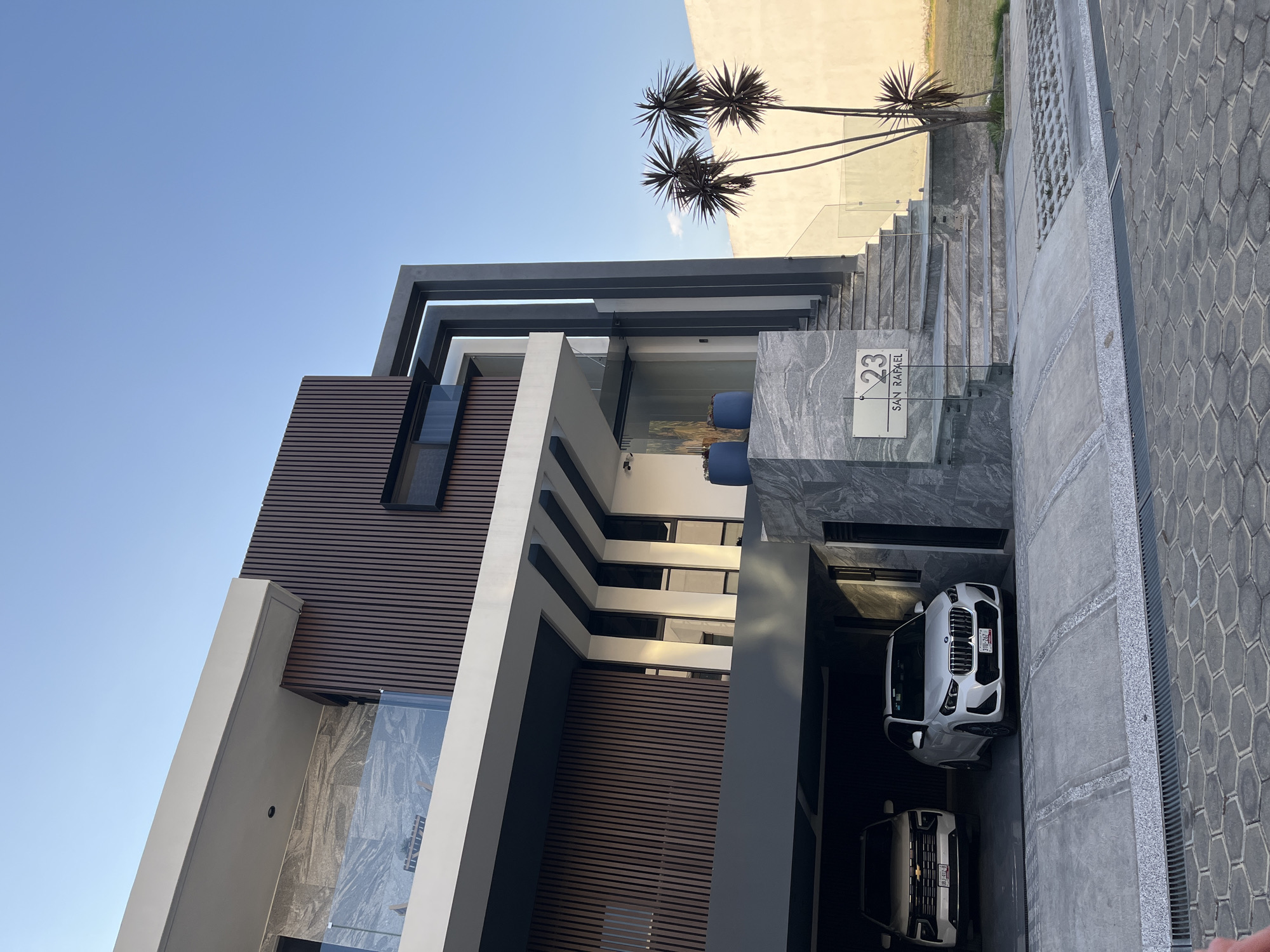 The photograph depicts a modern residence located at 23 San Rafael in San Andrés Cholula, Mexico. The architecture features a combination of materials, including dark brown horizontal wood slats, white and gray concrete framing, and glass railings. The building is multistory with visible windows. Two parked white vehicles occupy the driveway area, one of which is a BMW SUV with Mexican license plates. A stone staircase leads up to the entrance, marked by a sign displaying the address. The environment includes a clear blue sky, three palm trees, and a light-colored wall in the background. The ground is paved with gray cobblestones. No people are visible in the image.