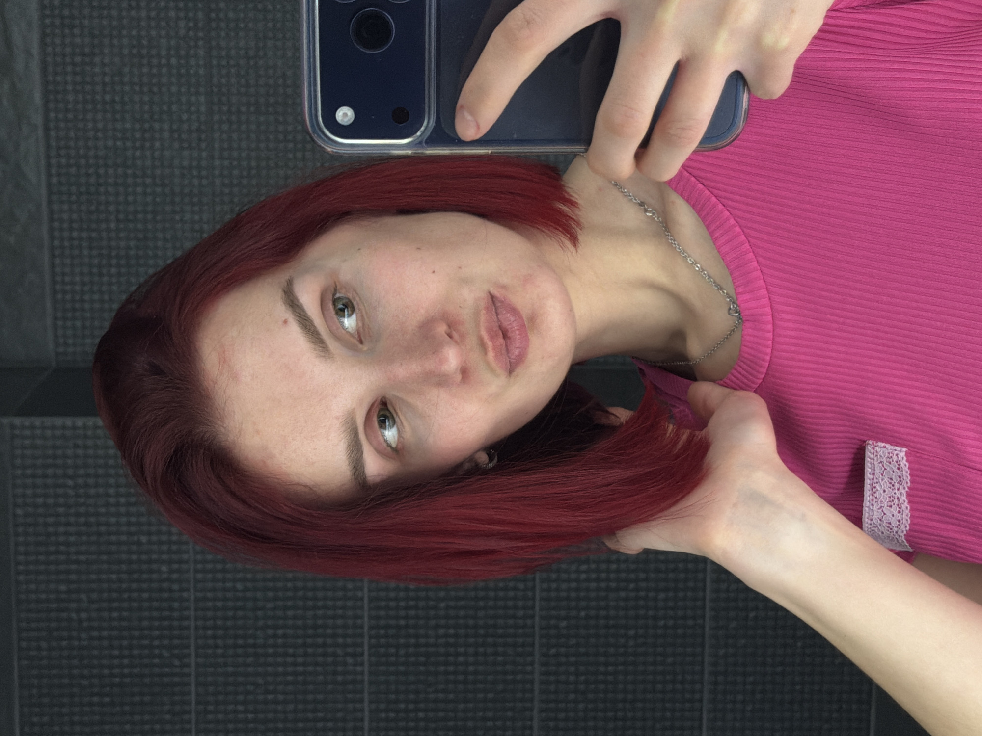 A young woman with red, shoulder-length hair is taking a selfie using her phone. She is wearing a pink shirt with a lace detail. The background appears to be a dark gray, tiled surface. The location is unspecified and cannot be determined. There is no indication it is in Chaikovsky, Russia.