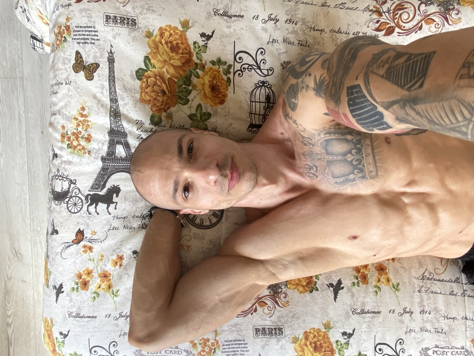 An adult male is pictured lying on his back on a bed, looking directly at the camera. He is shirtless, revealing a muscular physique and extensive dark tattoos covering both arms and his chest. On his right upper arm, the numbers "1960" are clearly visible. He has very short hair or is bald and a slight stubble on his chin, with a neutral to slightly smirking expression.
The setting is an indoor space, likely a bedroom, characterized by the patterned bedsheets he is lying on. The bedding features a Paris-themed design with illustrations of the Eiffel Tower, yellow-orange roses, butterflies, birds, a birdcage, and a vintage horse-drawn carriage. On the left side of the image, a light wooden floor is partially visible, indicating the floor surface next to the bed.
The man appears to be resting or posing casually. The lighting suggests it is daytime indoors, though the specific time of day or external weather cannot be determined. The actual location of this scene is Pleven, Bulgaria, despite the Parisian motif on the bedding.
Numerous texts are visible on the bedsheets, including: "PARIS," "Love Never Fails," "Colllutionne 18 July 1914," "things, endures all things," "POST CARD," "You," and a longer block of text stating, "This set contains 25 phrases to complement your kitchen and cusine themed projects. They are high quality clear." Other partially visible words include "Spadenascelle," "Kreebel," "Ze," and the number "13."