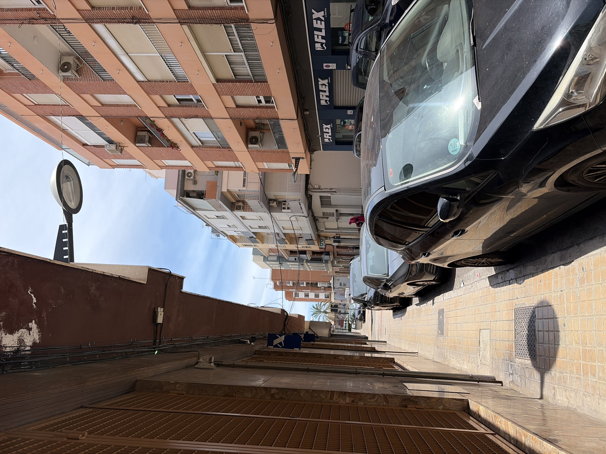 The image shows a street in Sagunto, Spain, with cars parked along the right side of the road. The street is lined with buildings on both sides; on the right, the buildings are taller, with some featuring balconies and window shutters. There is a 'FLEX' sign visible on one of the buildings. The building on the left is shorter and has a brown facade. A street lamp is also on the left side of the street. A palm tree is visible further down the street. The sun is shining, casting shadows from the streetlamp and cars onto the pavement.