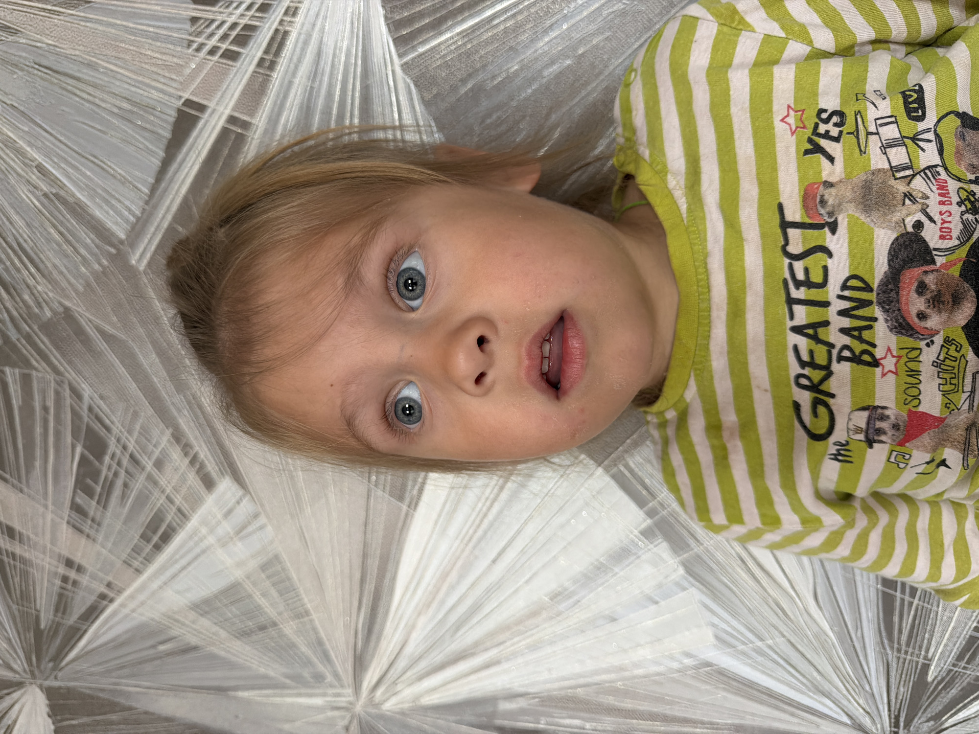 The image is a close-up portrait of a young child, likely a toddler, with blonde hair and blue eyes. The child is looking upwards and to the left with a surprised or curious expression, mouth slightly open. They are wearing a green and white striped long-sleeved shirt that features a graphic with meerkats and the text "GREATEST BAND." The background is a textured, reflective surface with a pattern of radiating lines, appearing to be wallpaper or a decorative panel. The lighting is soft, highlighting the child's face. The photograph was taken indoors, possibly in a residential setting in Chaikovsky, Russia, given the provided location context.