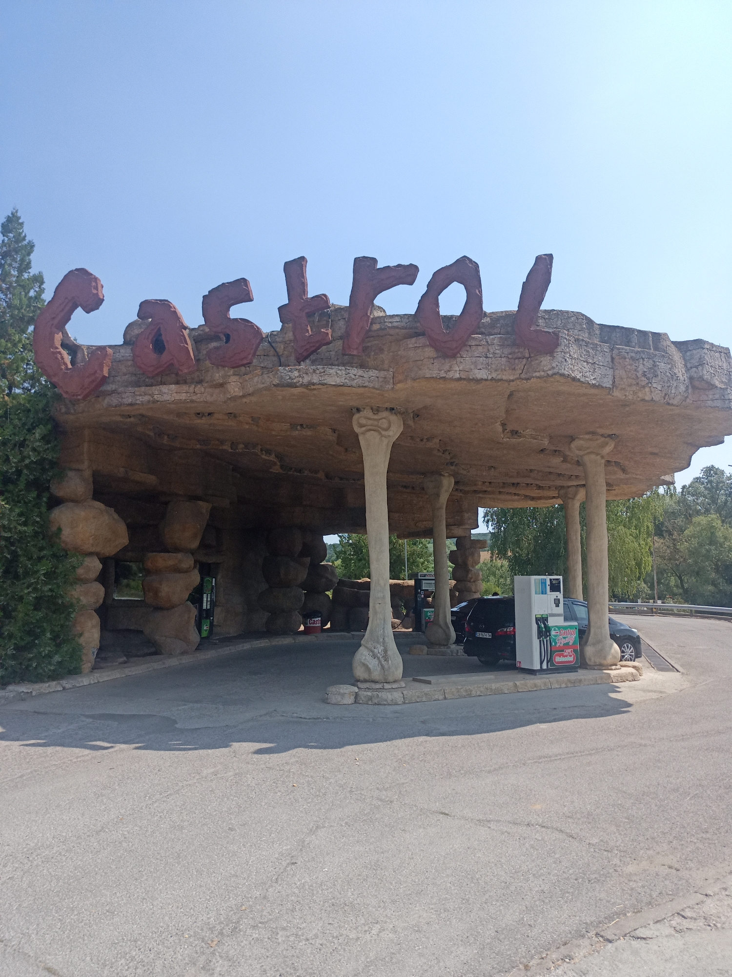 The image shows a Castrol branded gas station with a unique, cave-like architectural design. Large, blocky, red letters spelling "Castrol" are prominently displayed on the overhang of the structure. The overhang itself is made of a rough, textured material resembling rock. The supporting pillars also have a stone-like appearance, some resembling large bones.
Beneath the overhang, there are two cars and several gas pumps. One car, a dark-colored sedan, is parked next to a gas pump. The gas pump has a screen and hoses. A red and white Castrol oil can is visible on the ground near one of the pillars. The ground surface is asphalt.
The environment appears to be outdoors, with a clear blue sky and some trees visible in the background. The overall impression is of a themed gas station, likely designed to evoke a prehistoric or naturalistic aesthetic.
This specific gas station is located in the city of Omurtag, Bulgaria, as indicated by the presence of a license plate on the parked car which appears to be Bulgarian.