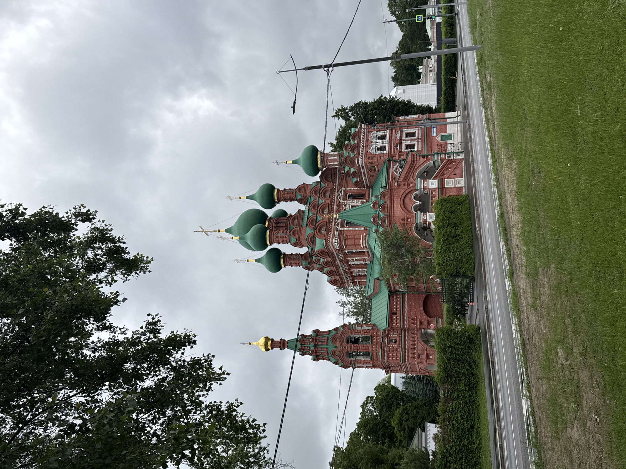 The image captures a red brick building with green onion domes, likely a church, under a cloudy sky. The building is located next to a road and a grassy area in what appears to be a somewhat urban setting. Vegetation, including trees and bushes, surrounds the structure, adding to the scene's overall ambiance. The photograph's location is Khimki, Russia.