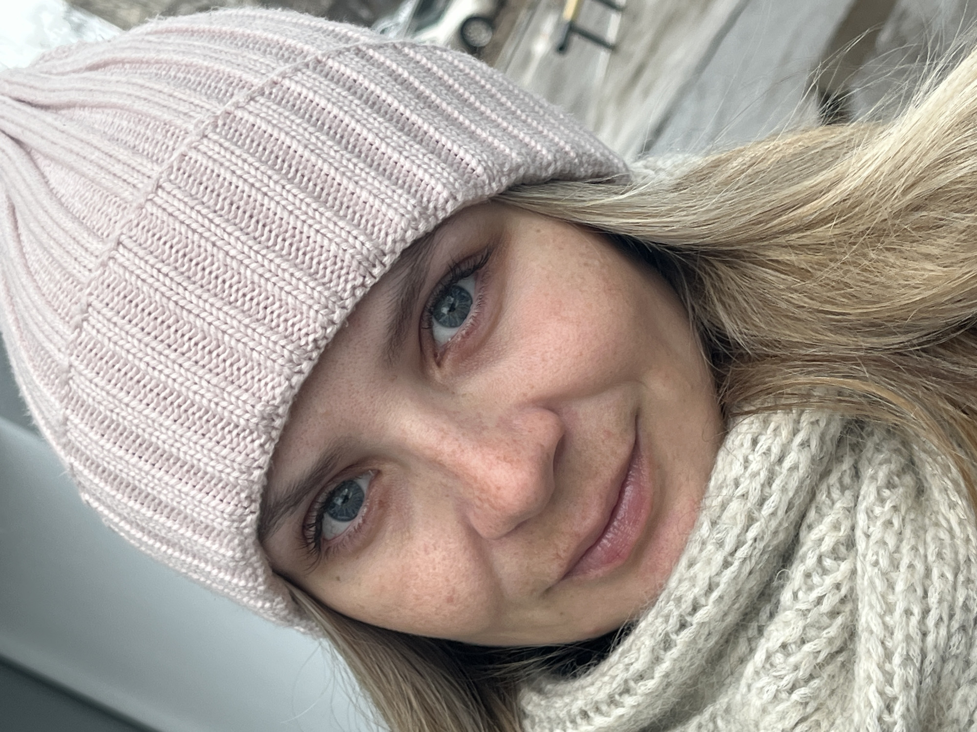 A close-up selfie of a woman with blonde hair, wearing a pink knitted beanie and a light gray knitted scarf. She has fair skin, blue eyes, and freckles. The background is blurred but appears to show a city street, with a car and street signs visible. While the image may be taken in a city, there is no information within the image to specifically confirm the location as Chaikovsky, Russia.