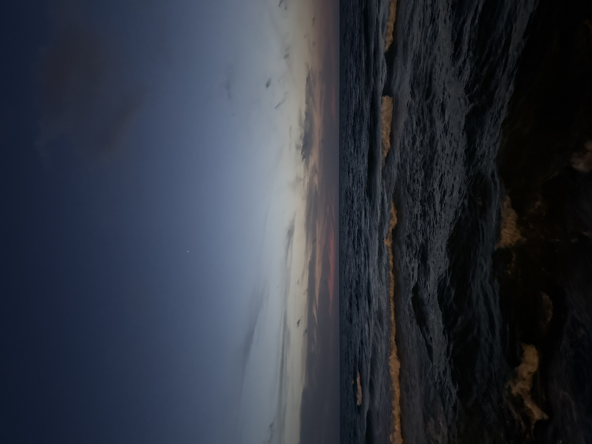 The image depicts a seascape at dusk, likely from Tel Aviv, Israel, although no specific landmarks confirm this. The sky is a gradient from dark blue to lighter hues with muted pinks and yellows near the horizon. There are scattered clouds. The sea is choppy with visible waves and dark-colored water. There is some light reflecting off the wave crests, producing a golden glow. No people or objects are visible on the beach.