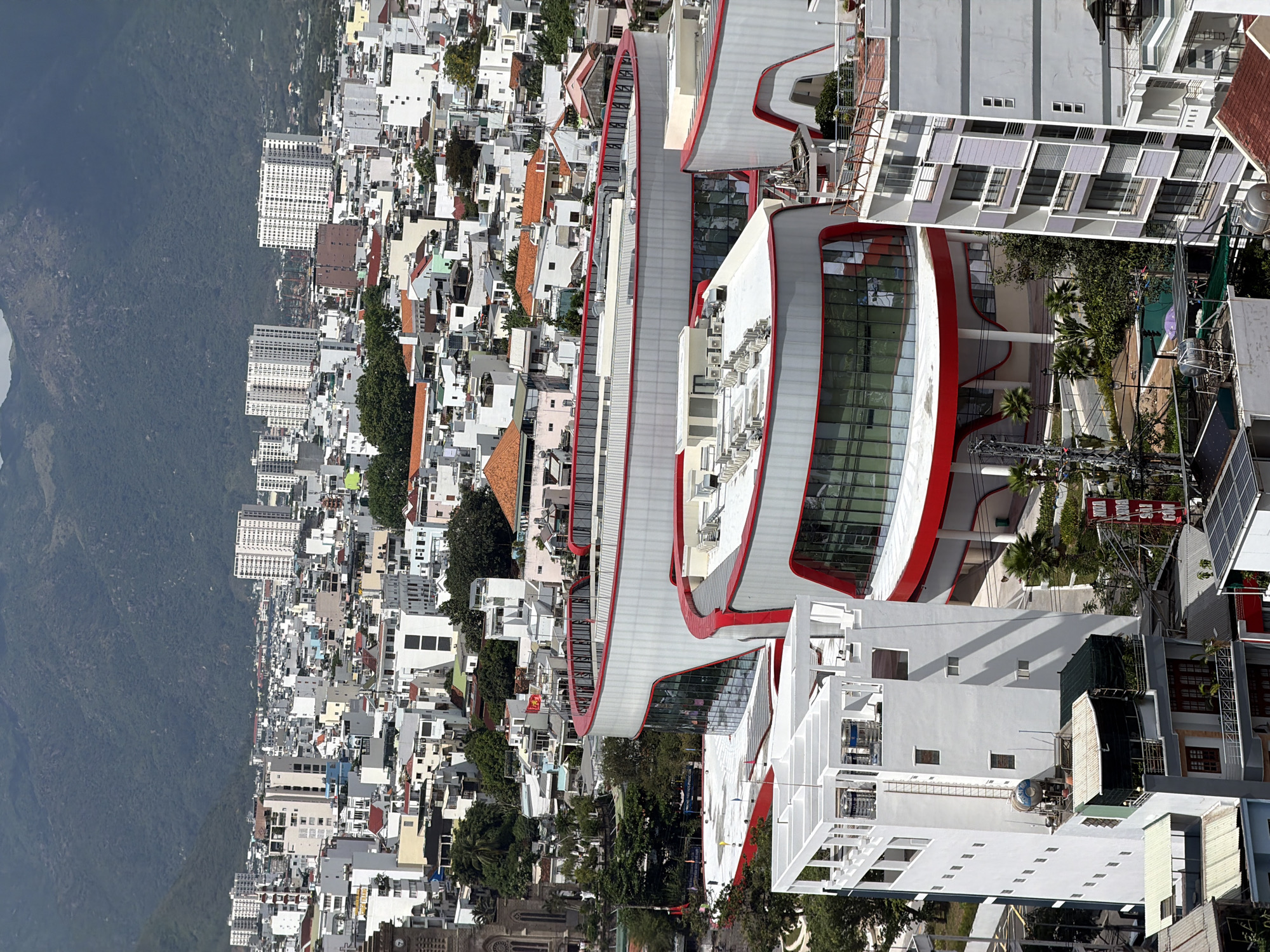 This high-angle, panoramic photograph showcases a vibrant urban landscape in Nha Trang, Vietnam, set against a backdrop of lush, forested mountains under bright daylight.
The city sprawls across the frame, characterized by a dense collection of residential and commercial buildings of varying heights and styles. In the mid-ground, a striking modern architectural complex dominates, featuring a distinctive curved design with prominent red and white accents and extensive glass facades. This multi-level structure contrasts sharply with the surrounding buildings, which are predominantly white, grey, and light-colored, many with flat or tiled roofs. Numerous taller, multi-story buildings, likely apartment blocks or hotels, are visible throughout the urban expanse, particularly further back towards the mountains.
The cityscape displays a high population density, with buildings packed closely together. Pockets of greenery, including palm trees, are interspersed among the structures. Essential urban infrastructure, such as power lines and poles, is visible in some areas, alongside solar panels on several rooftops.
The weather appears sunny and clear, casting distinct shadows across the buildings. A Vietnamese flag, red with a yellow star, is visible on a flagpole amidst the buildings, confirming the location. Inside one of the modern buildings, a sign with "188 BAR" is faintly discernible. The image captures the bustling appearance of Nha Trang on a regular day, with no specific activity or event taking place beyond general urban life.