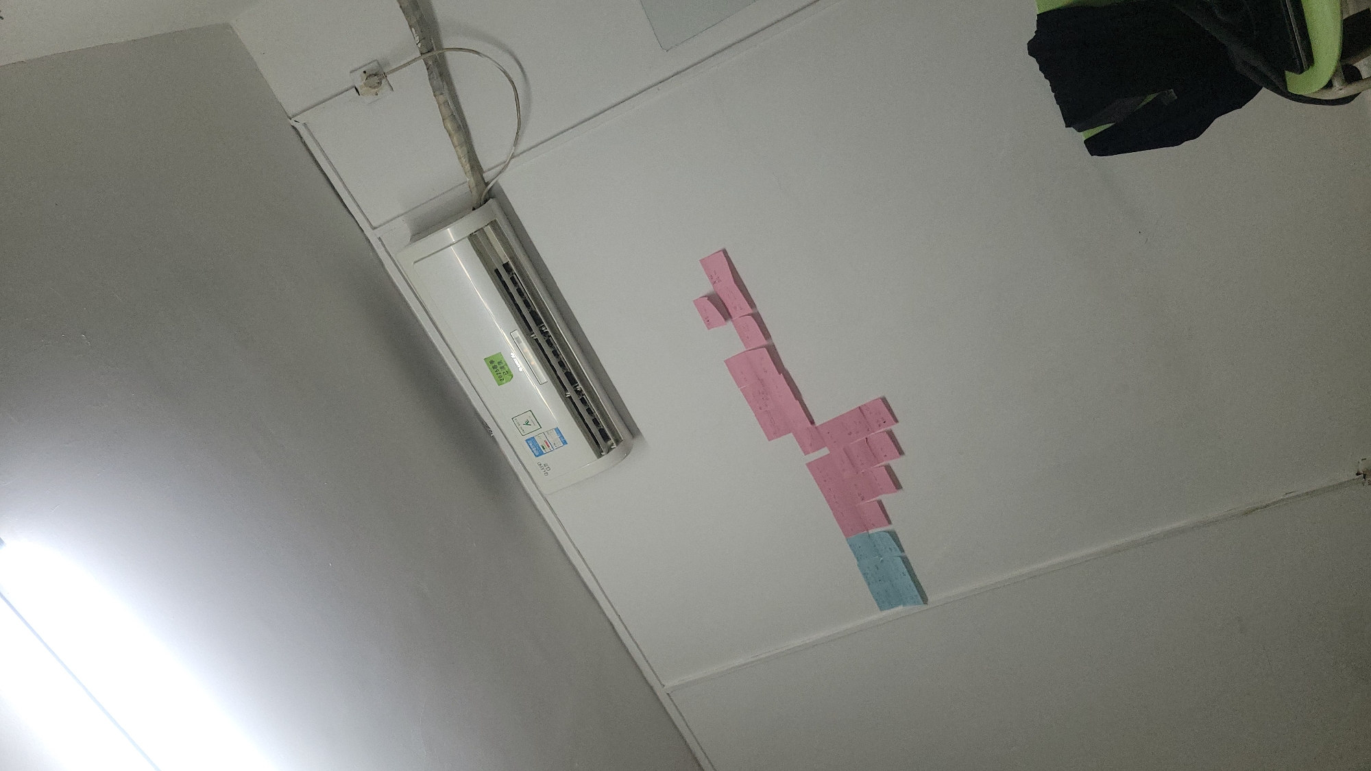 A photograph taken from a low angle captures a section of a room's ceiling. The ceiling is predominantly white with an air conditioning unit mounted on the left side. To the right of the air conditioner, a series of pink and blue sticky notes are arranged in a descending pattern. In the upper right corner, a piece of dark fabric or clothing is visible. The photograph shows an indoor environment with artificial lighting, presumably located in Yuzhong District, China, though no geographical information is discernible in the visual data.