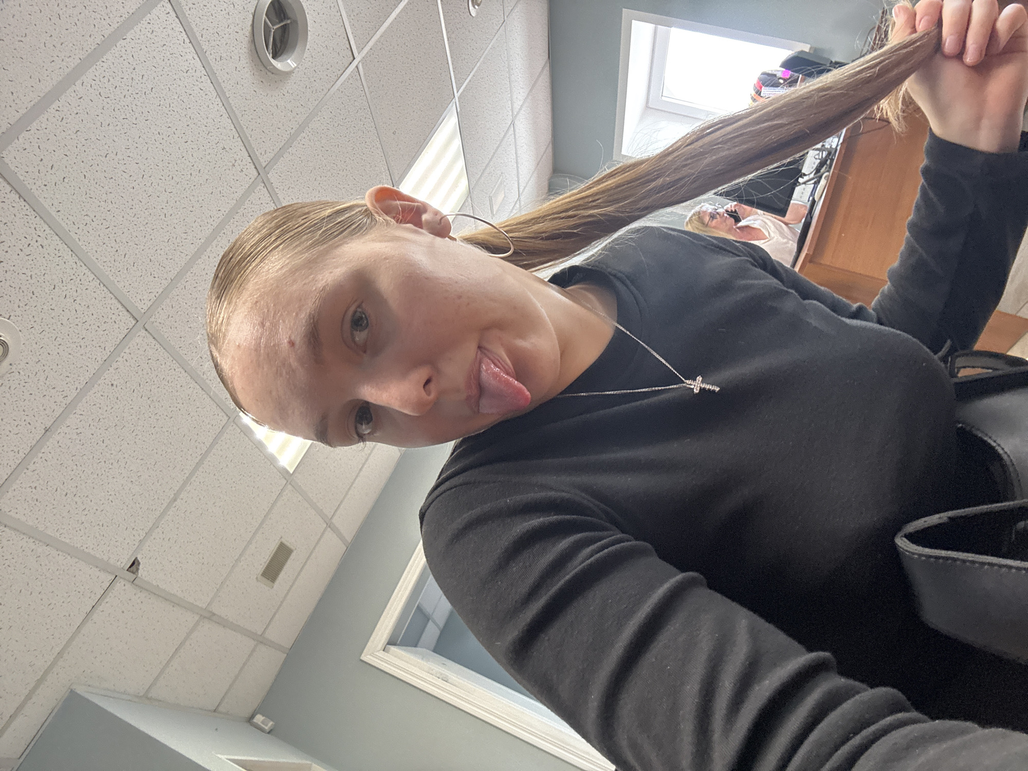 The image shows a young woman with blonde hair pulled back in a ponytail. She is wearing a black long-sleeved shirt and a necklace with a cross pendant. She is sticking her tongue out and looking at the camera. The background appears to be an office with a dropped ceiling, fluorescent lighting, and a doorway leading to another room. A woman in the background is sitting at a desk and talking on the phone. The scene is indoors, likely in an office or administrative building. There is no indication of the city of Perm, Russia in the image.