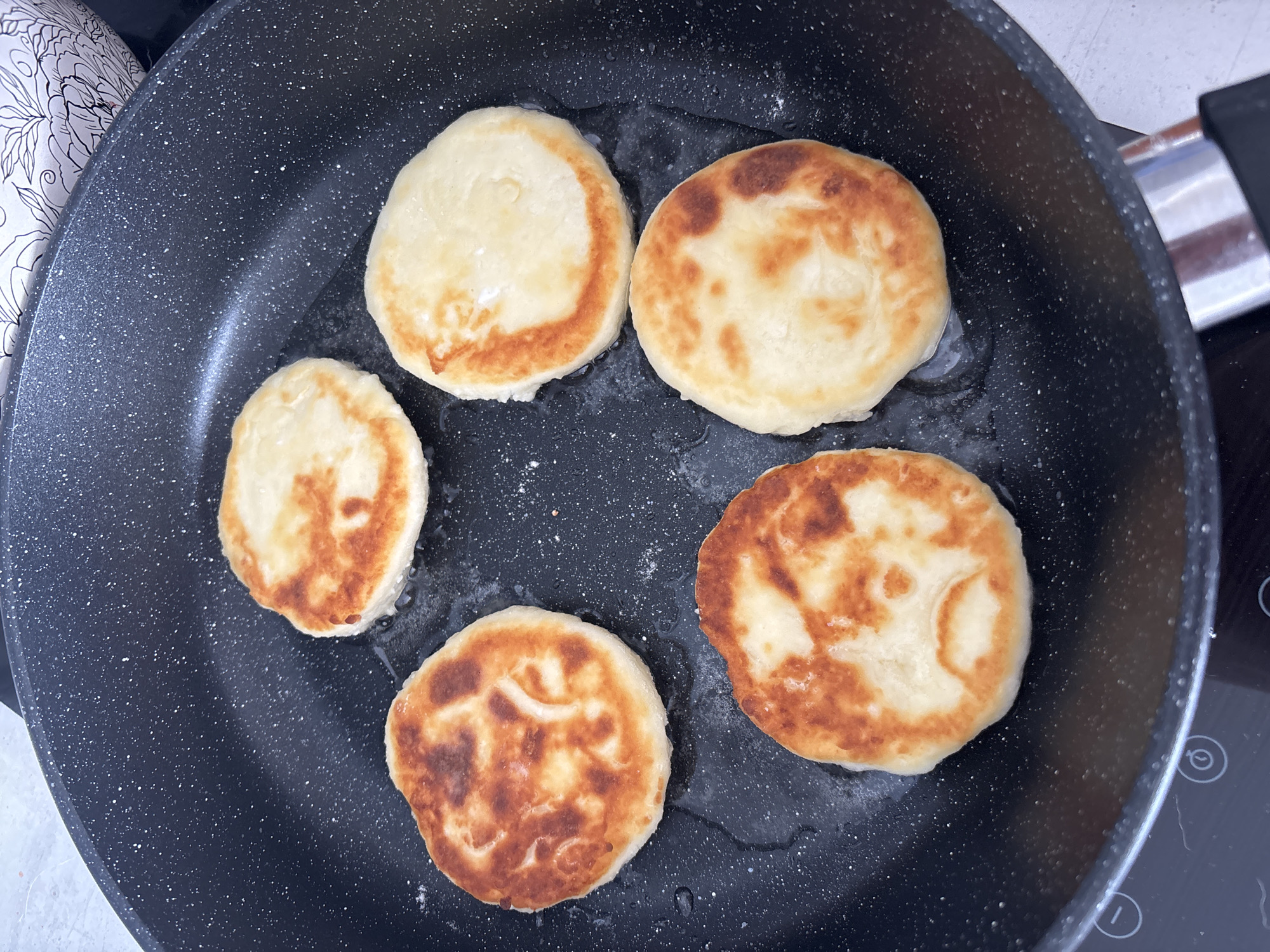 The image shows six circular, dough-based food items frying in a black non-stick pan with white speckles. The items display various levels of golden-brown caramelization on their surfaces, indicating they are in the process of being cooked. A thin layer of cooking oil is visible in the pan. A portion of a black induction stovetop control panel, featuring white etched symbols including a timer icon and plus/minus buttons, is visible in the lower left. In the upper right background, a segment of a white ceramic or porcelain object with black floral or leafy patterns is partially visible. No individuals or direct interactions are present. The indoor environment lacks any specific visual cues that would enable the identification of its geographic location, including the city of Zmeinyi, Russia.