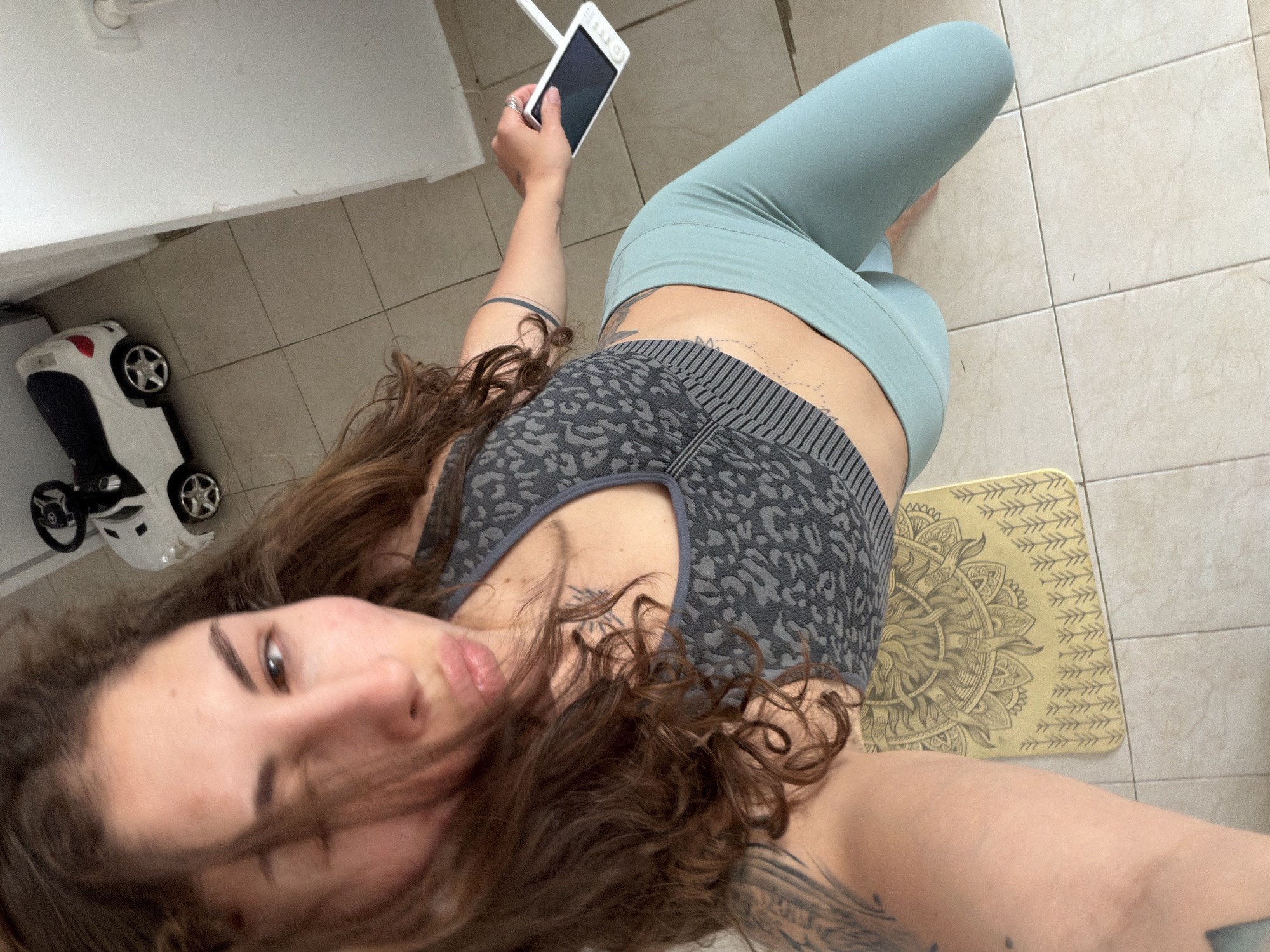 A woman is taking a selfie indoors. She is lying on a yellow yoga mat on a tiled floor. She is wearing a grey patterned sports bra and light blue leggings. She is holding a white smartphone in her left hand. There is a white toy car near a white wall in the background. The location is potentially Kiryat Bialik, Israel.