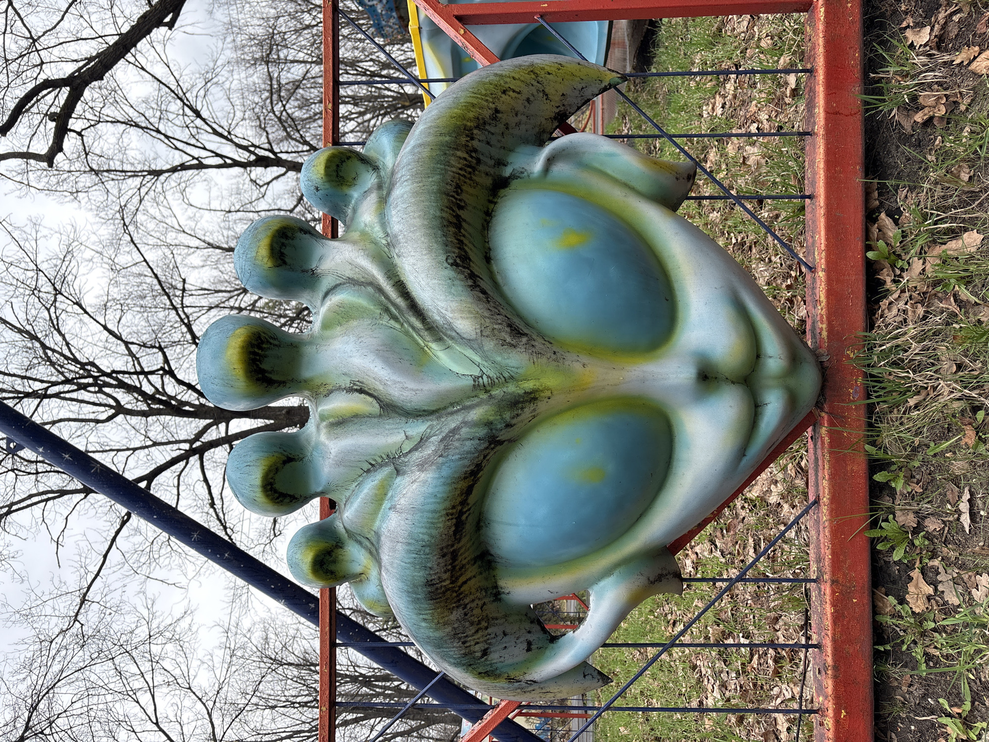 A large, light blue and green, mask-like sculpture resembling a fantastical sea creature with oversized blue eyes is mounted on a red and blue metal frame outdoors. The sculpture is detailed with swirling patterns and bulbous shapes protruding from one side. The frame is set against a backdrop of brown, dry leaves and patches of green grass. Bare tree branches are visible in the background, suggesting a park or outdoor recreational area. The location is an unknown area of the city of Ulyanovsk, Russia.