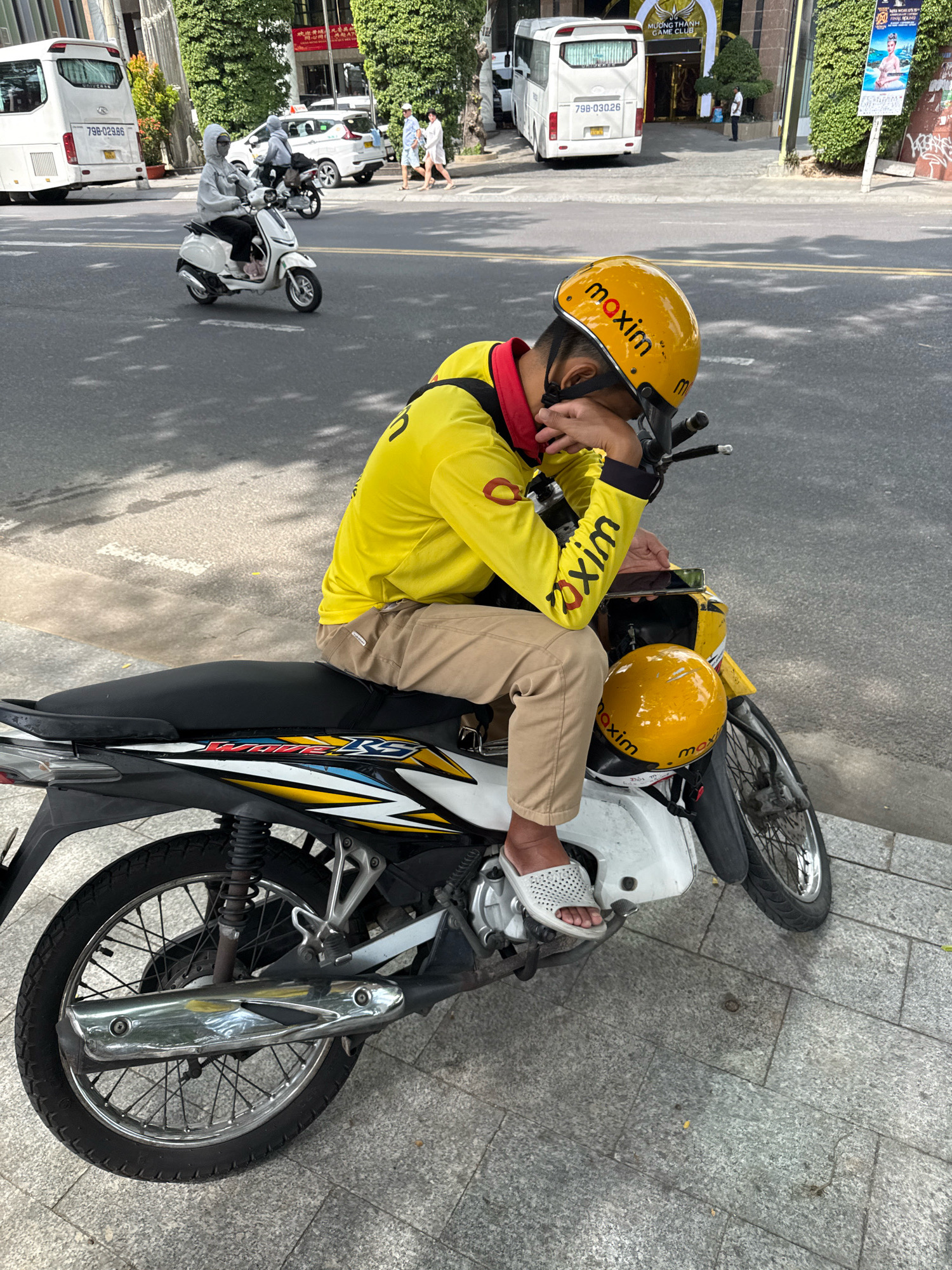 A person wearing a yellow uniform with "Maxim" branding is resting their head on their hand while sitting on a white motorcycle. The motorcycle has "Maxim" logos on its yellow helmet and the person's uniform. The person is wearing khaki pants and white slide sandals. The background shows a street scene in Nha Trang, Vietnam, with trees, buildings, and other vehicles, including a white bus and a white scooter. The person appears to be taking a break or is tired.