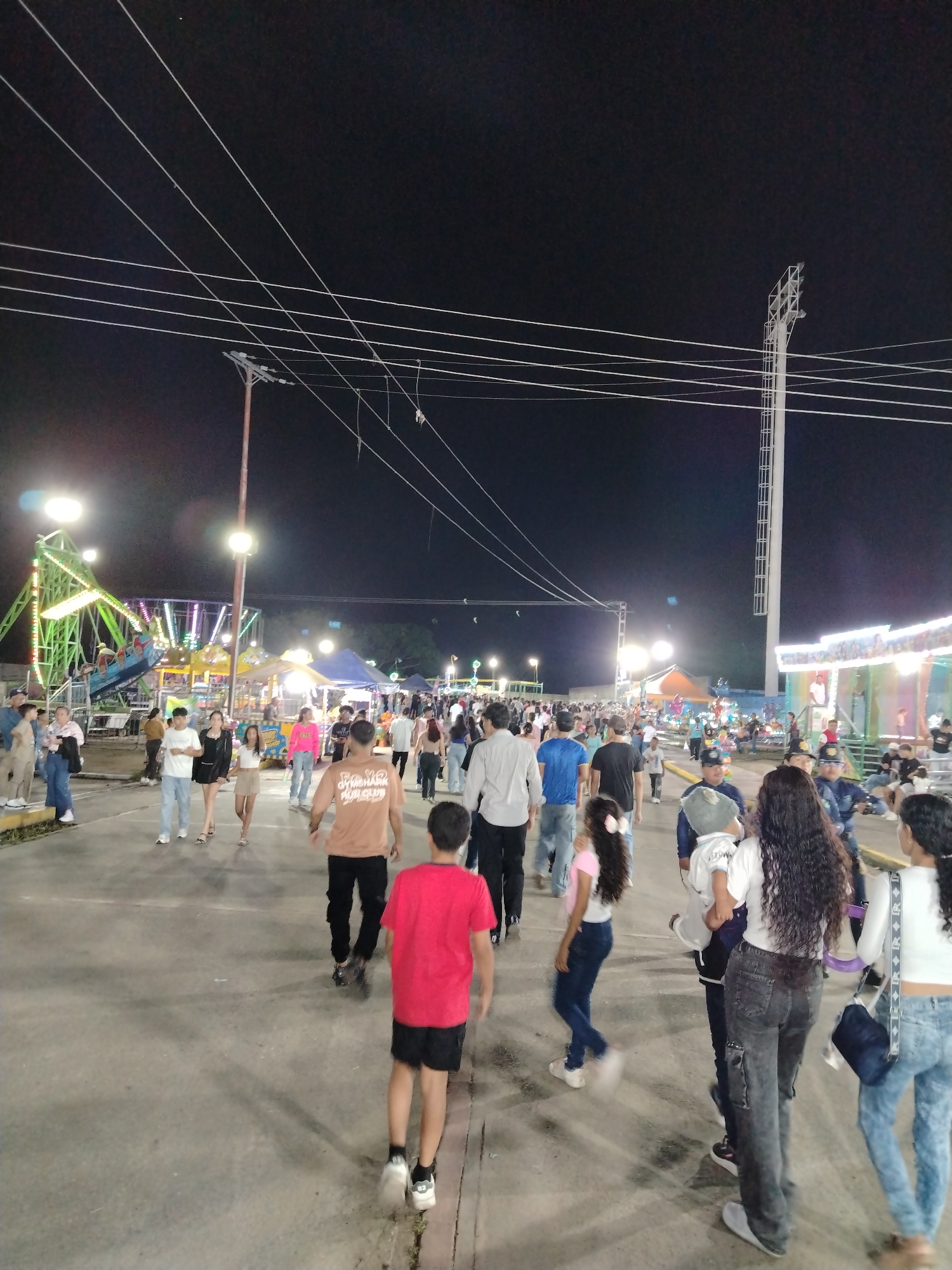 This media file depicts a lively outdoor fair or carnival scene at night in Tinaquillo, Venezuela. Numerous people, including adults and children, are seen walking along a paved thoroughfare, bustling through the event.
The setting is characterized by a dark night sky, illuminated by a multitude of artificial lights emanating from various fair attractions, stalls, and tall light poles. Overhead, a complex web of white power lines spans the frame, connected to both wooden and tall metal utility poles.
In the midground on the left, a brightly lit green swing ride is prominently visible, with people on board. Further into the fair, other illuminated rides and tents with colorful lights add to the festive atmosphere. On the right side of the path, more fair stalls and structures are present.
The crowd consists of a mix of individuals and groups, all casually dressed, moving in various directions along the pathway. Their relaxed posture suggests a pleasant and engaging experience. A notable detail is a man in the immediate foreground, walking away from the camera, wearing a tan t-shirt with the visible text "GYMSHARK RUN CLUB" printed on the back. The ground is a light-colored paved surface, reflecting some of the ambient light. The overall scene conveys a vibrant community gathering enjoying a recreational event under the night sky.
