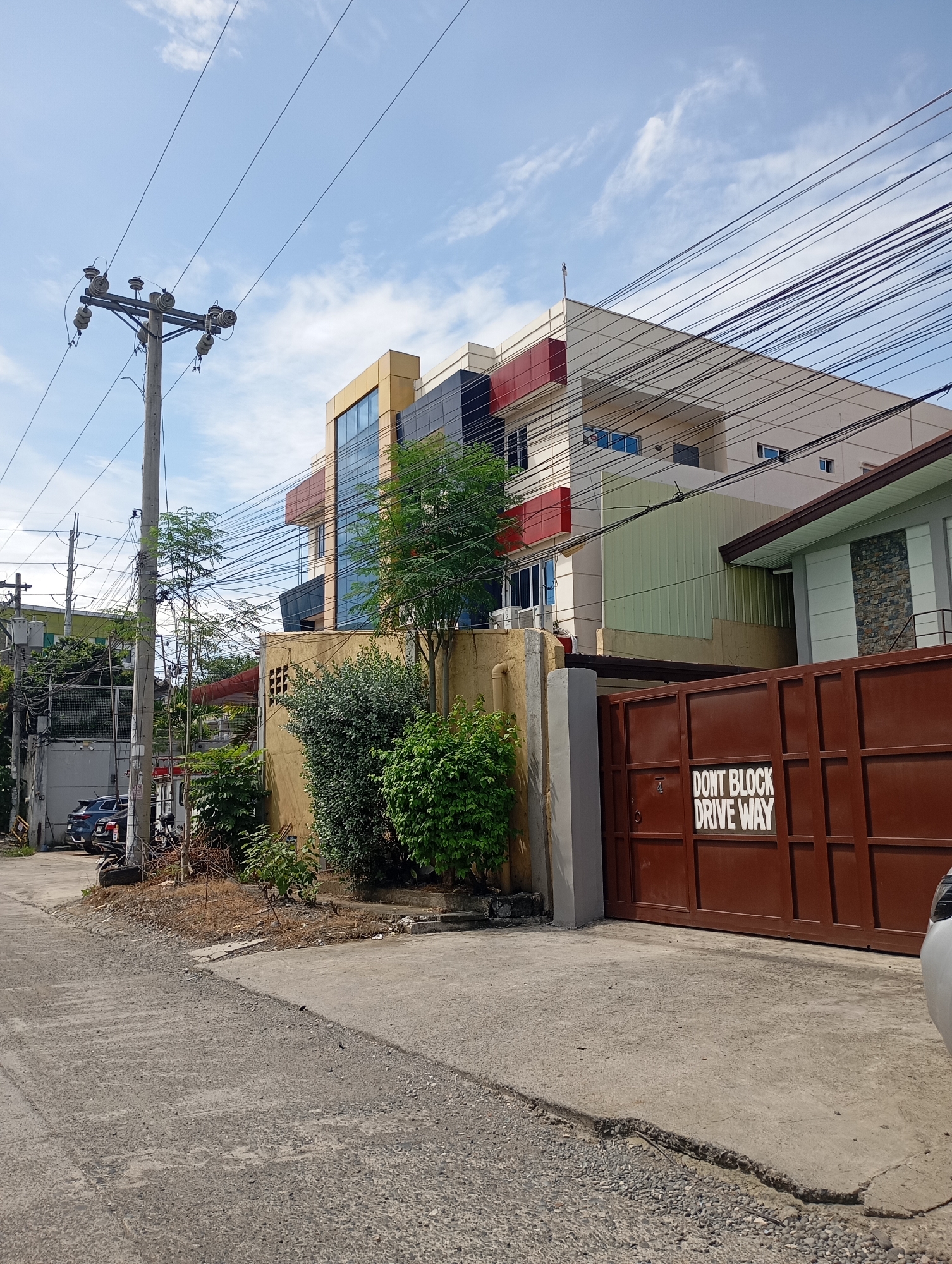 This daytime image captures a street scene in Davao City, Philippines, under a bright blue sky dotted with scattered white clouds. The foreground features a concrete road or driveway, transitioning from a rougher texture to a smoother surface. On the right, a prominent dark brown metal gate is partially visible, bearing the white text "DON'T BLOCK DRIVE WAY" and the number "4." A small portion of a white car is also seen at the extreme right edge of the frame.
A tall wooden utility pole, heavily laden with numerous overhead electrical and communication wires, dominates the left side of the midground. These wires stretch across the sky, creating a complex web. Behind the gate and along the street, lush green bushes and small trees thrive, adding natural elements to the urban landscape.
The background reveals a cluster of buildings with varied architectural styles. A modern, multi-story building stands out with its facade incorporating large glass panels alongside sections of yellow, red, and black. Adjacent to it is a lighter-colored building featuring a section with decorative stone cladding and a dark-tiled roof overhang. Further down the street on the left, additional buildings are visible, with two parked cars, including a blue SUV and a motorcycle, suggesting a mix of residential and commercial activity. The scene is devoid of people, presenting a quiet snapshot of an urban environment characterized by its dense utility infrastructure and tropical foliage.
