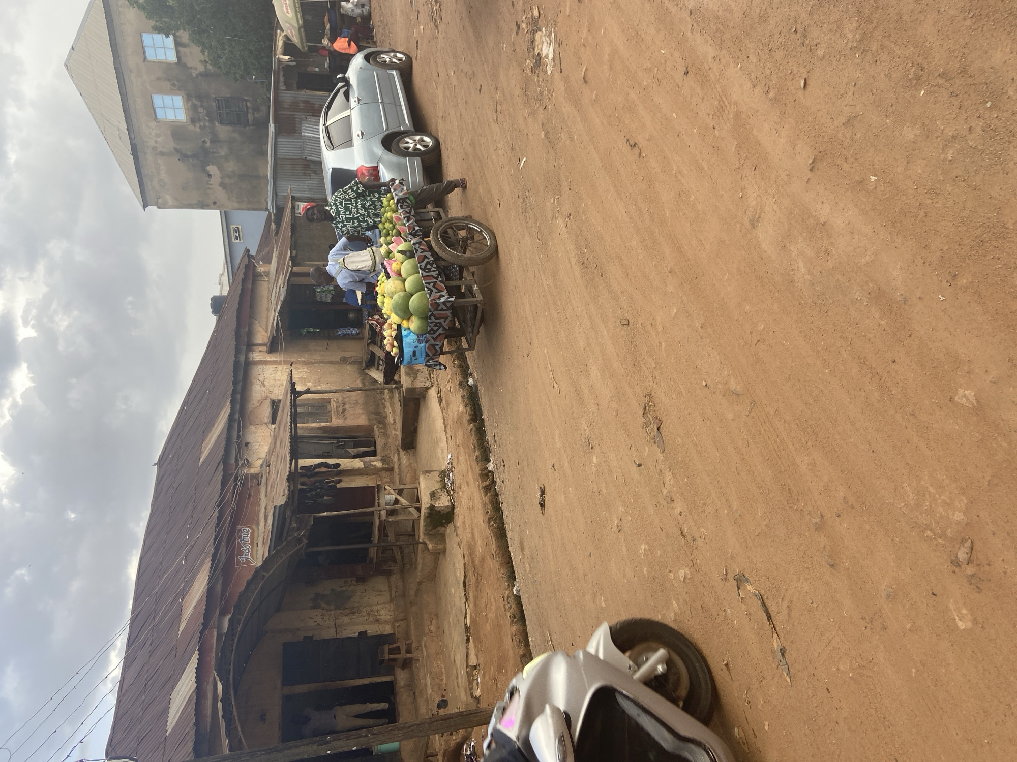 In Bukuru, Nigeria, a dusty street scene unfolds. A light blue sedan is parked alongside a weathered, corrugated metal-roofed building. In front of the building, a fruit vendor pushes a cart laden with green watermelons and other produce. Two men stand near the cart, one wearing a white shirt and backpack, the other in a green patterned shirt. A silver motorcycle is partially visible on the left side of the frame, suggesting ongoing activity on the street. The environment is characterized by the unpaved, reddish-brown earth of the road, interspersed with scattered debris.