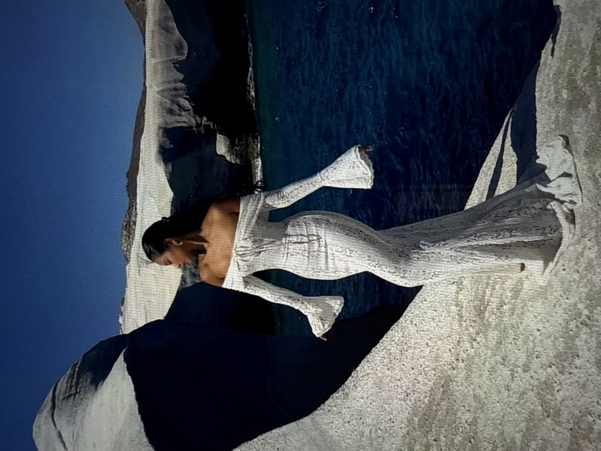 A woman stands on a rocky shore, wearing a long, white, off-the-shoulder dress with flared sleeves and a lace overlay. She is looking out at the bright blue sea. Behind her, white rock formations rise from the water, with a clear blue sky overhead. The scene is bathed in bright sunlight. The location is not specified as Găvănoasa, Moldova.