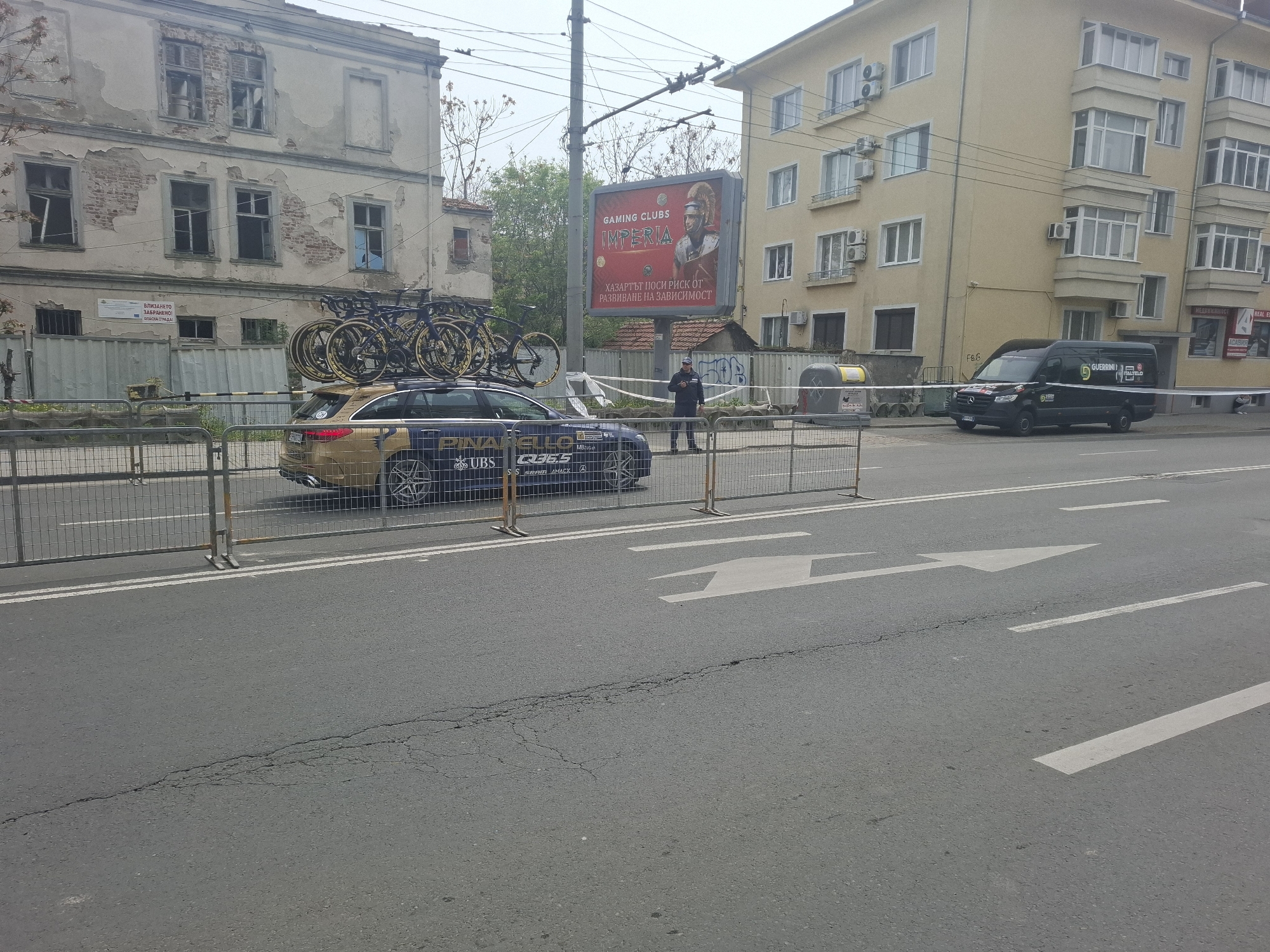 The image depicts a street scene in Burgas, Bulgaria. A gold-colored station wagon with multiple bicycles strapped to its roof is parked behind metal barriers. The car has "Pinarello," "UBS" and "Q36.5" logos on its side. A man in a dark uniform stands near the car. Behind the car, a billboard advertises "Gaming Clubs Imperia" with a warning about the risk of gambling addiction. To the right of the billboard is a yellow apartment building. A black van with "Guerrini" and "FBC" logos is parked across the street. A dilapidated building with visible brickwork is visible behind the car. The road has white lane markings and a white arrow indicating direction.