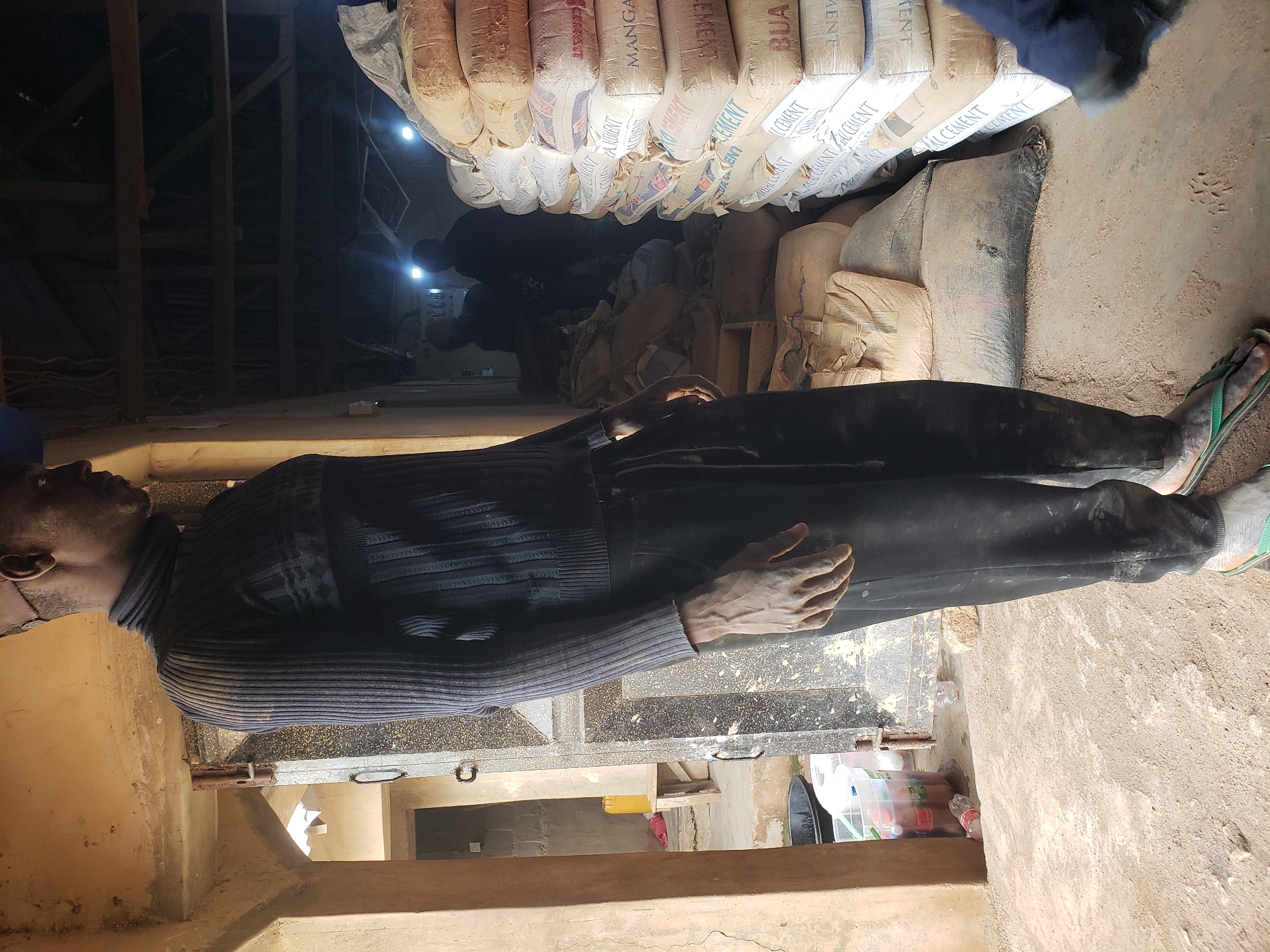 The image depicts a man standing in a dimly lit, dusty environment, possibly a storage area or workshop in Jos, Nigeria. He is wearing a dark, ribbed turtleneck sweater and dark pants, with his feet clad in green flip-flops. The man is seen from the waist down, with his left hand resting on his thigh.
The background reveals stacks of cement bags, some with visible branding like "BUA CEMENT" and "MANGANESE CEMENT". The floor is covered in dirt and fine dust, suggesting an outdoor or semi-outdoor setting. In the far background, two figures are dimly visible, silhouetted against illuminated areas, implying activity within the space. To the left, a doorway leads to another area with a yellow container and a wooden bench, further indicating a functional, possibly industrial or construction-related location within Jos. The overall impression is one of a work environment, characterized by its rough, dusty nature and the presence of building materials.