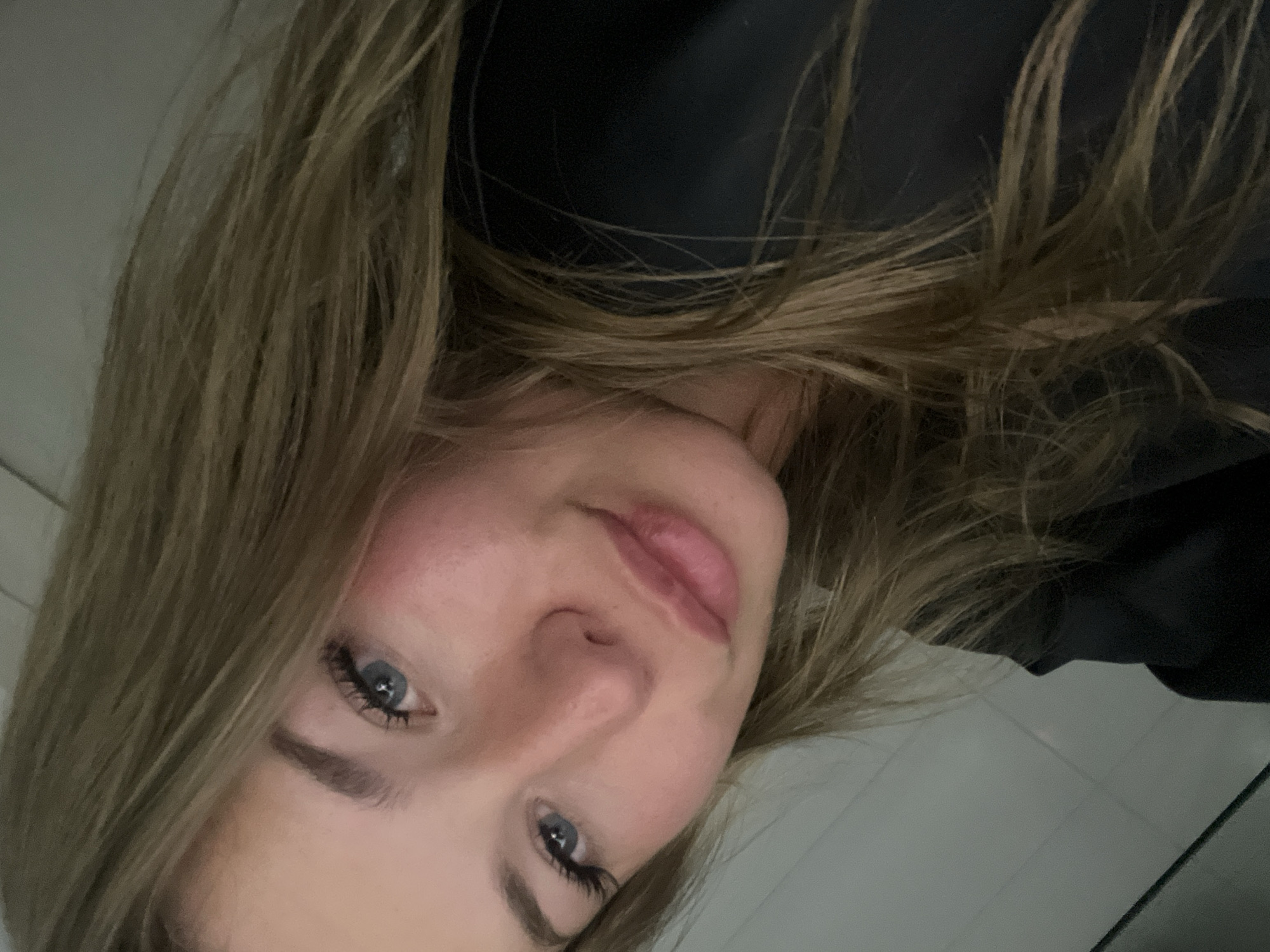 A close-up, rotated selfie photograph depicts a young, fair-skinned female with shoulder-length blonde hair. She has blue eyes, visible eyelashes, and pink lips. She is wearing a dark-colored top. The background consists of a white tiled surface and a dark-colored object. The location, Budva, Montenegro, cannot be determined based solely on the given visual information.