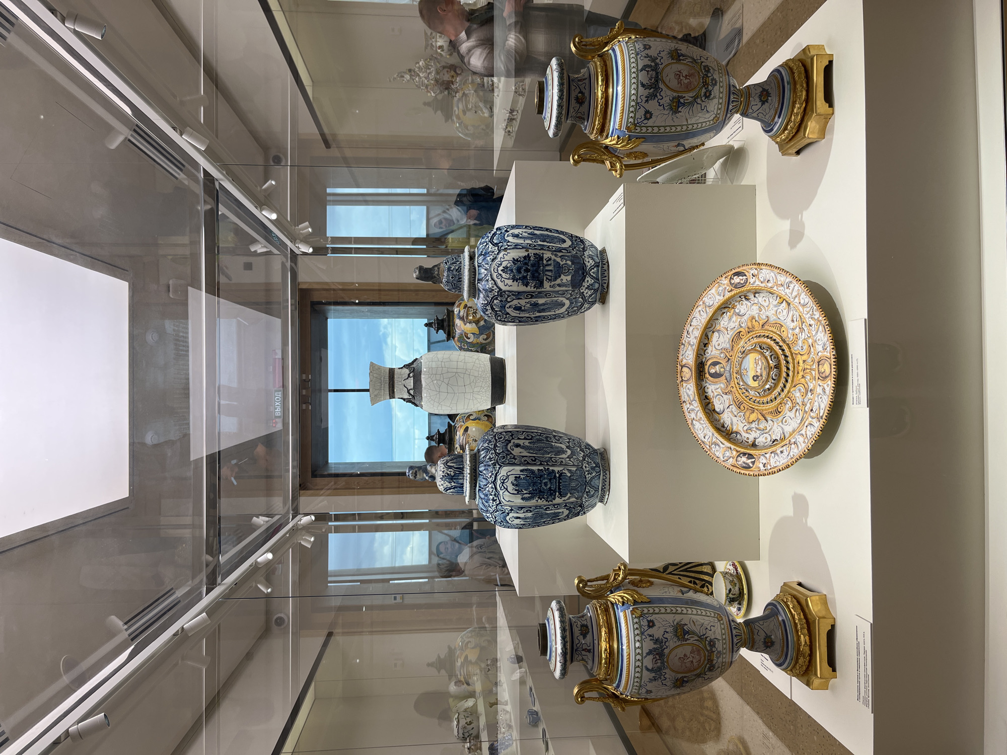 The image shows a display case in a museum, filled with antique porcelain vases and a plate. The vases are primarily blue and white, with some featuring gold accents and ornate handles. A plate with a central image surrounded by gold and other patterns is also present. The display is lit from above, and the objects are arranged on white platforms within the glass case. The location appears to be indoors, possibly a museum or gallery setting in the city of Unknown, Unknown. Reflected in the glass of the display case are silhouettes of people and the window behind them.