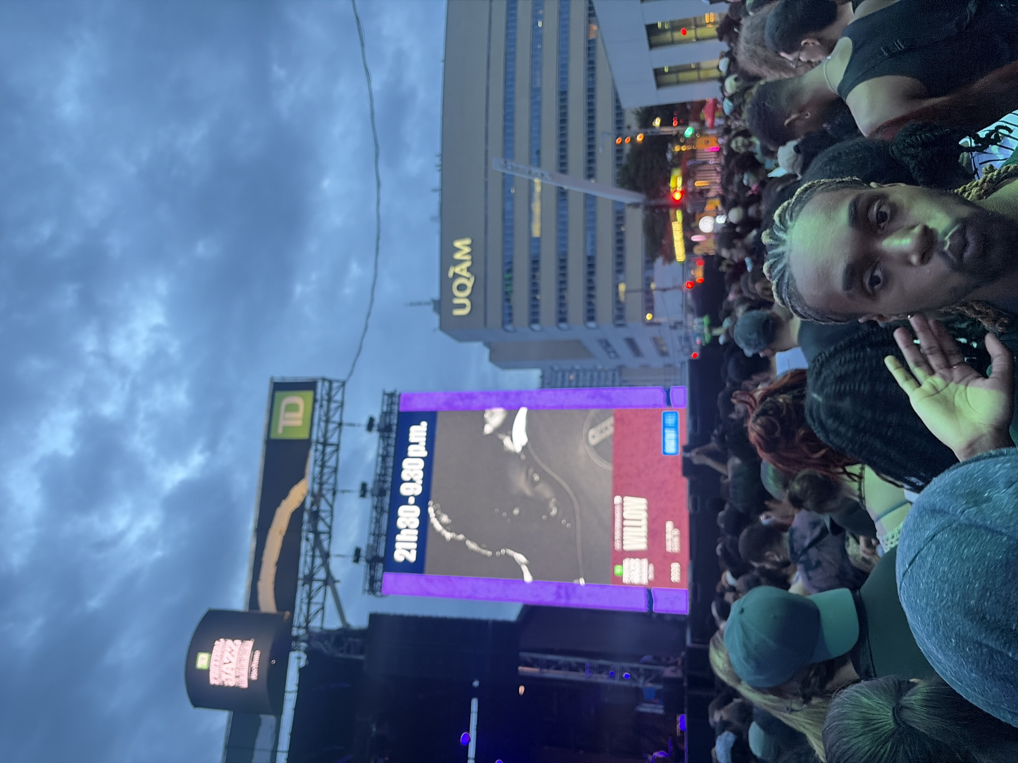 This image captures an outdoor evening event in Montreal, Canada, likely a music festival given the large stage, screens, and crowd. The sky is overcast and dark blue, suggesting twilight or evening.
In the background, a large digital screen displays "21h30-21h30 p.m." and an image of a person's face, possibly an artist named Willow, as indicated by text below. Above this screen, banners with the TD logo and "Festival Jazz" are visible, further confirming a festival setting. To the right, the "UQAM" building is prominent, identifying the location as near the Université du Québec à Montréal. Traffic lights and illuminated signs indicate a city street setting adjacent to the event space.
The foreground is filled with a diverse crowd of people attending the event. Many are looking towards the stage or the screens. One man in the immediate foreground is taking a selfie, with his right hand raised near his face. The atmosphere appears to be lively and engaged, with people gathered closely together.
