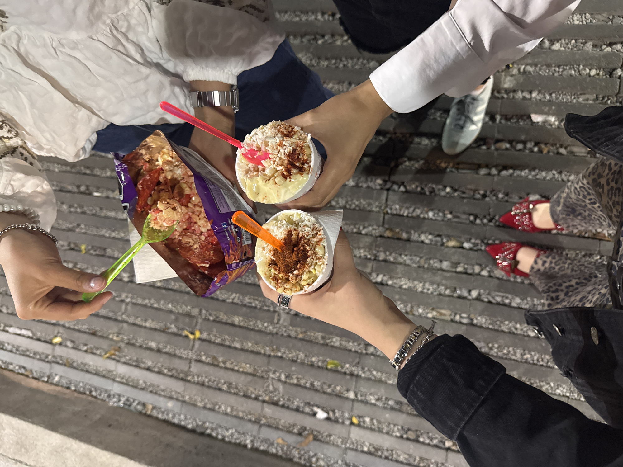 Four individuals are partially depicted from an overhead perspective, focusing on their hands, arms, and lower bodies, gathered around street food. The scene takes place outdoors on a paved surface composed of dark grey bricks or patterned concrete, with some scattered yellowish debris, suggesting an urban setting in Mexico City during the evening or night.
The central activity involves people holding and consuming Mexican street snacks. Two hands hold white cups filled with what appears to be esquites—a Mexican street corn salad—topped with white cheese and generous amounts of red chili powder, each served with a bright plastic spoon (one red, one orange). Another hand holds a large, purple snack bag, which has been cut open and filled with a variety of toppings, resembling Tostilocos. These toppings include corn kernels, red sauced items (possibly cueritos or pickled pork rinds), and other spices, served with a neon green plastic spoon. The word "Doritos" is partially visible on the purple bag.
The attire of the individuals includes a white, ruffled blouse with a silver watch, a plain white long-sleeved shirt, and a black denim jacket adorned with silver bracelets and a ring. Footwear visible includes light blue sneakers and distinctive red, sparkly flat shoes worn with leopard-print pants. The overall impression is one of casual, social snacking.