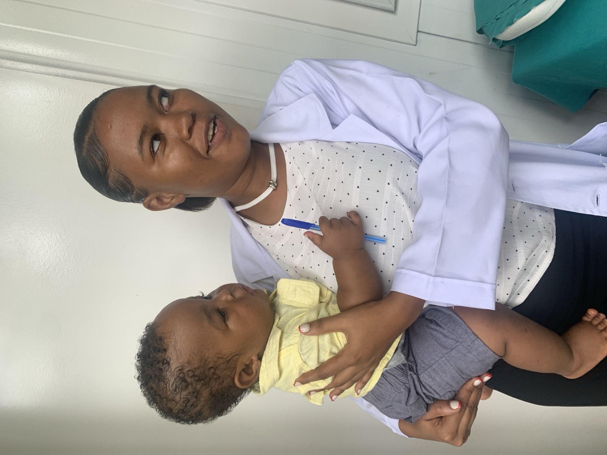 An adult female holds an infant in an indoor setting in Dar es-Salaam, Tanzania. The female, positioned on the left side of the frame, wears a white lab coat over a white polka-dot blouse and a white choker necklace with a silver-colored charm. Her dark hair is pulled back with styled edges, and she looks towards her right (viewer's left) with her mouth slightly open. Her right hand holds a blue pen, resting on the infant's torso, while her left hand supports the infant's back. Her fingernails feature a white tip and red accent manicure. The infant, held securely in the female's arms, wears a yellow collared shirt and grey shorts, with bare feet visible. The infant has dark, curly hair and looks in the same general direction as the female. The background consists of a white textured wall or door paneling. To the right of the female, green fabric and a white cushion are partially visible.
