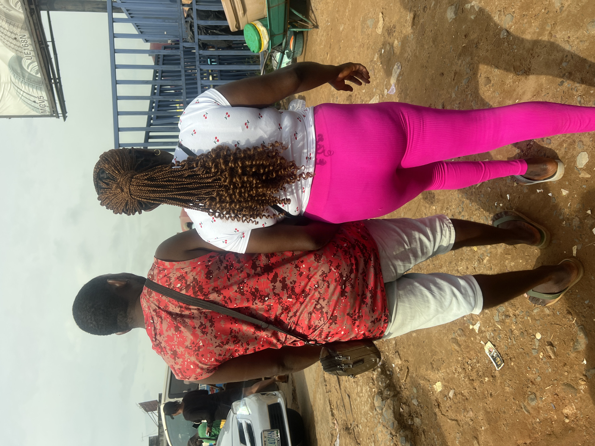 In Onitsha, Nigeria, two individuals walk away from the camera on a dirt path. The person on the left is a man wearing a sleeveless red shirt with a pixelated camouflage pattern in red and black, and light grey shorts. He has a dark brown bag slung across his chest. The person on the right is a woman wearing a white t-shirt with small red cherry prints and bright pink leggings. Her hair is styled in numerous thin braids that form a ponytail. Behind them, a blue metal fence encloses an area with some large industrial containers and a green wheelbarrow. A billboard with tire advertisements is visible in the background, and a white car is parked to the left. The ground is uneven and covered in dirt, gravel, and scattered debris.