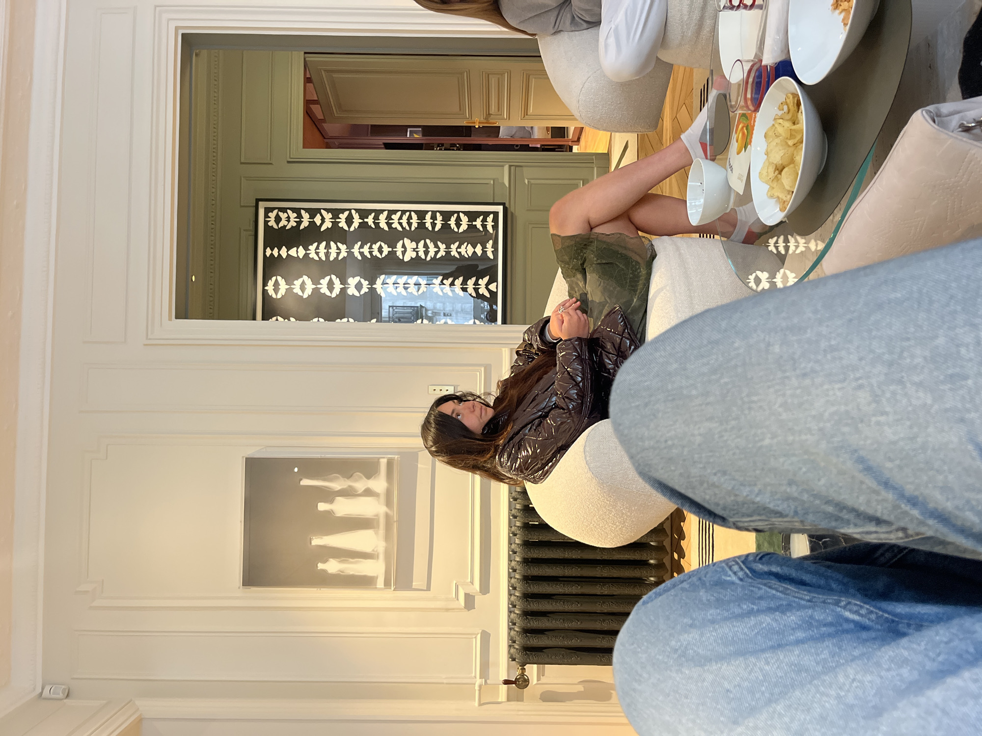 A medium indoor shot shows a woman sitting on a chair next to a radiator, while another person in blue jeans is visible in the foreground. The woman is wearing a brown, shiny jacket and is looking to the left. A framed black and white artwork is behind her, and another framed print is on the wall behind the radiator. A doorway leads to another room, where a black-and-white picture is visible, as well as part of a sofa. On a table next to the sofa, there are snacks in bowls. The setting appears to be a stylish apartment or lounge, possibly in Paris, France, based on the architectural details.