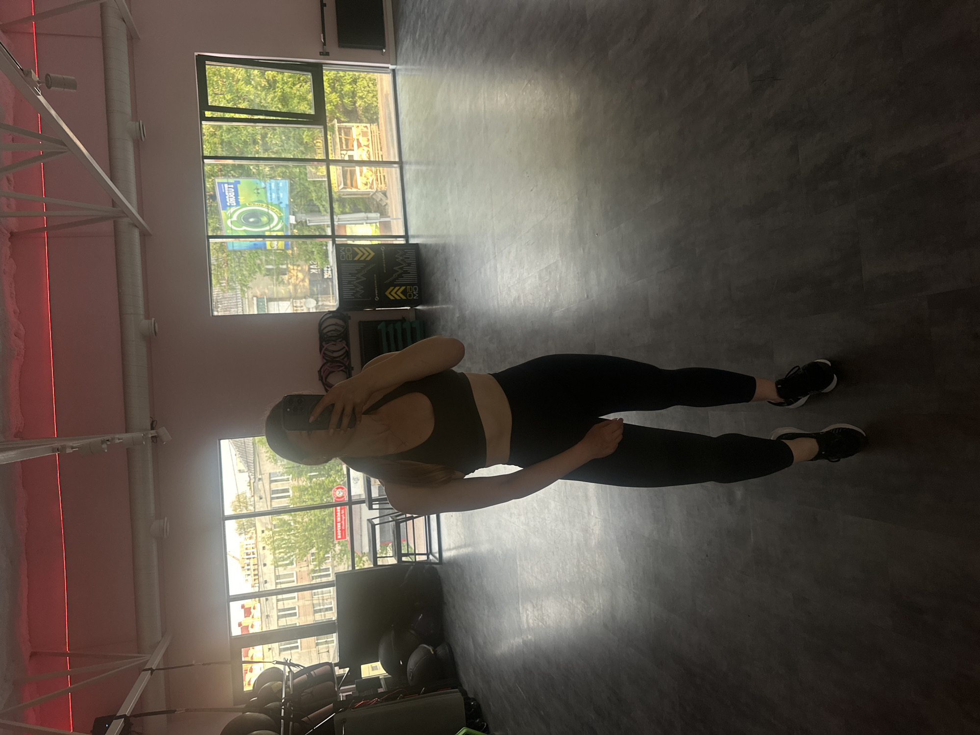 A mirror selfie captures a person standing in what appears to be a fitness studio or gym. The subject, a person with light-colored hair, is positioned centrally, holding a smartphone to take the photo. They are wearing dark athletic attire, consisting of a sports bra or crop top and leggings, paired with black sneakers featuring white soles.
The studio features a dark gray, textured floor and light pink walls. Exposed white structural elements and ventilation ducts run across the ceiling, with red strip lighting providing an accent along one wall. Large, multi-pane windows occupy one side of the room, allowing ample natural light to enter and offering a view of the outside.
Through the windows, green trees and urban buildings are visible, suggesting a city setting during the daytime. Several signs are seen through or reflected in the windows. One prominent blue and green sign clearly displays the Cyrillic word "СПОРТ" (Sport) at the top, followed by "ТОБІ МОЛОДЬ!" (To you, youth!). Another red sign, visible in reflection, shows partial Cyrillic text that appears to start with "БАРИ" (Bari).
The overall impression is a modern gym space, likely in Ternopil, Ukraine, given the Cyrillic text on the external signage. The activity captured is a casual self-portrait, possibly before or after a workout.