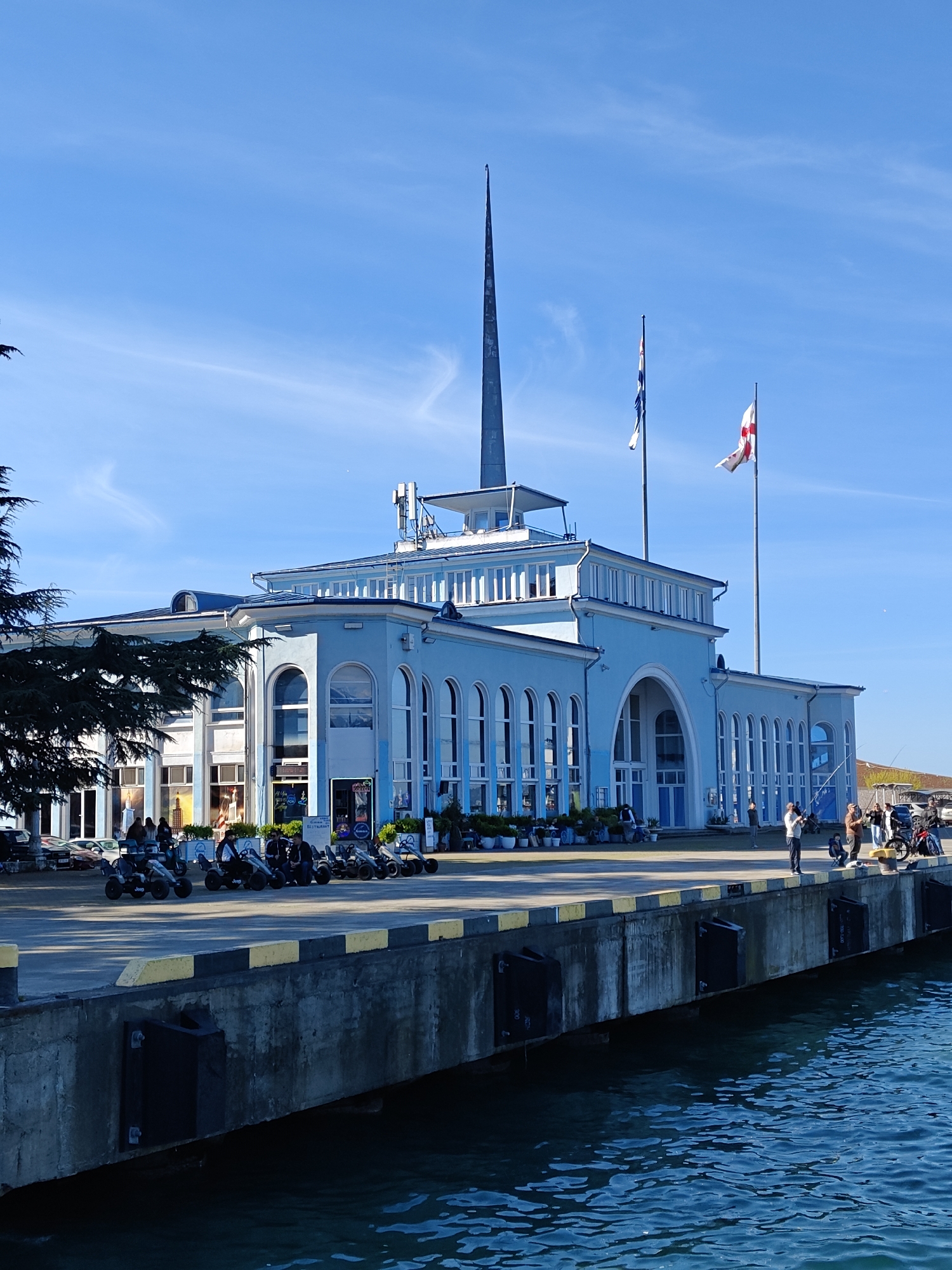 The image shows the Batumi Sea Port in Batumi, Georgia, under a bright blue sky. The port terminal is a light blue building with a central tower topped by a spire, flanked by two flagpoles flying the flags of Georgia and Adjara. A row of arched windows extends along the front of the building. A sidewalk runs in front of the building, and people are visible both in front of the building and along the waterfront. Several pedal-powered jeeps are parked on the sidewalk, and a group of people is fishing along the waterfront. The water is a deep blue with visible ripples. The foreground includes a concrete pier with black rubber bumpers.