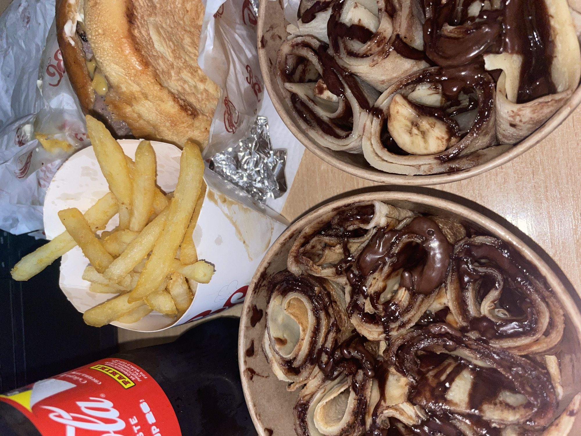 The image shows an assortment of fast food items arranged on a surface, likely a table. The items include a partially eaten burger in wrapping, a serving of french fries in a paper container, two bowls filled with rolled pancakes topped with chocolate syrup and what appear to be banana slices, and a bottle of Coca-Cola, only partially visible at the bottom left corner. The background is plain and does not provide any clues about the location or the city of Plovdiv, Bulgaria. There is no additional data to substantiate a city or location-specific description. The focus is on the food items.