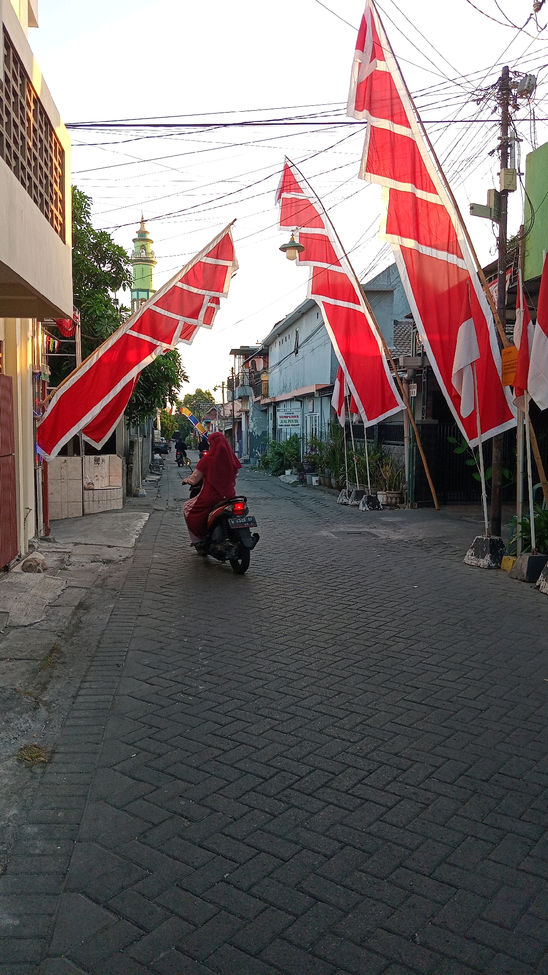 In the city of Yosowilangun, Indonesia, a person wearing a red hijab and dress is riding a motorcycle down a narrow, brick-paved street. The rider is facing away from the camera. Red and white flags, likely Indonesian flags, are prominently displayed on poles along the right side of the street and also in the foreground, extending across the street. The flags are large and billow in the breeze. On the left side of the street, a building with a patterned facade is visible. In the background, a green minaret of a mosque can be seen rising above the rooftops of other buildings. Power lines are strung overhead, typical of a populated area. The scene appears to be set during daylight, possibly late afternoon or early morning, given the lighting.