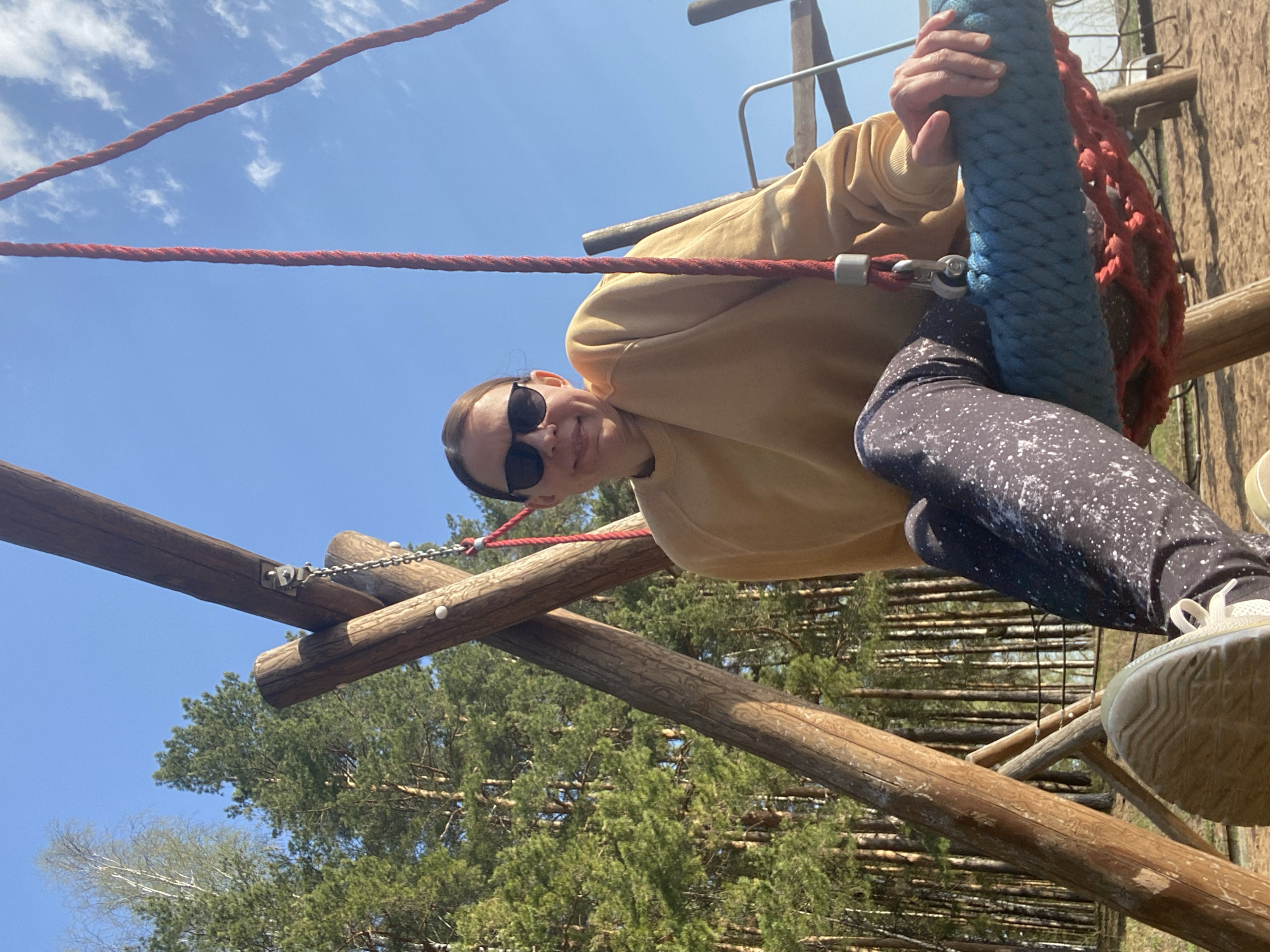 A woman wearing sunglasses and a light yellow sweater is on a rope course in a forest in Chaikovsky, Russia. She is gripping a thick blue rope with a red net around it. She's wearing dark leggings with white speckles and tan sneakers. The rope course is made of wooden logs and red rope, and the trees are visible in the background against a blue sky.