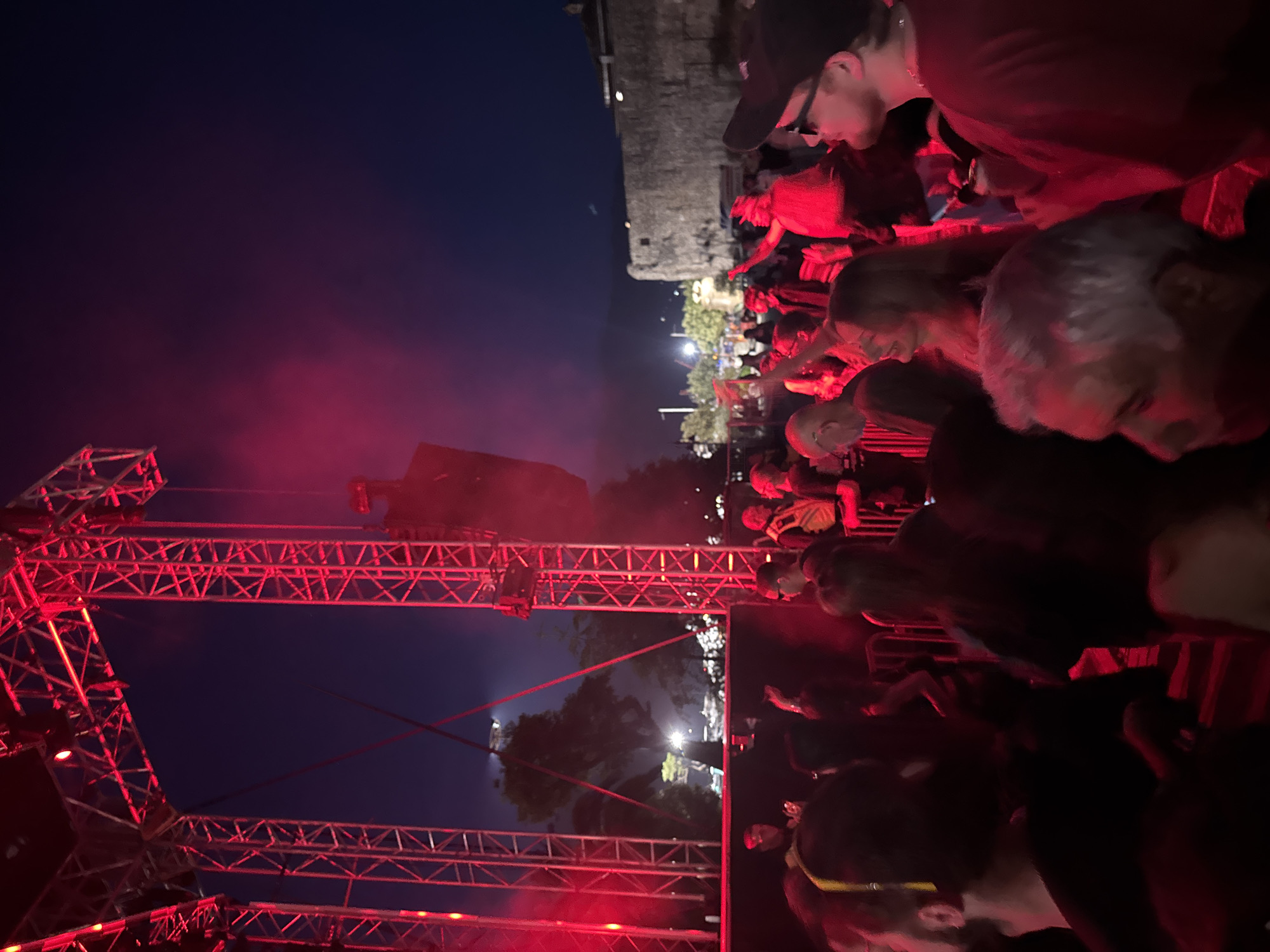The image captures an outdoor concert in Budva, Montenegro, at night. A large crowd is gathered, illuminated by red stage lights. A metal stage truss is visible on the left side of the image, also lit in red. Above the crowd in the background are streetlights and an old stone building. The sky is dark blue. People are standing in close proximity, looking towards the stage.