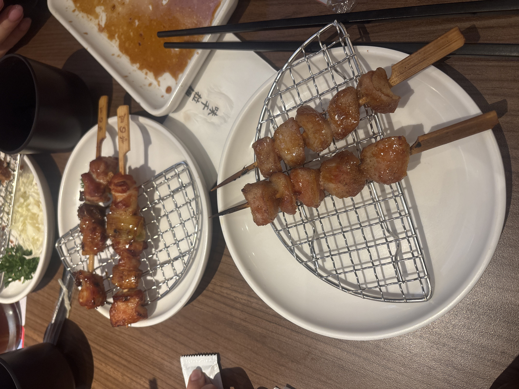 The image depicts a table setting with several plates of food and drinks, likely in a restaurant. In the foreground, a white plate holds two skewers of cooked meat, possibly chicken or pork, resting on a small metal rack. The meat is glazed and appears to be slightly charred. Another white plate is visible in the upper portion of the image, also containing skewers of similar meat. Adjacent to the plates, a dark mug and a partially consumed dish with sauce are present. Two black chopsticks lie on a white napkin with Asian characters. The table surface is made of wood. The scene suggests a meal in progress. The image location is Haizhu District, China.